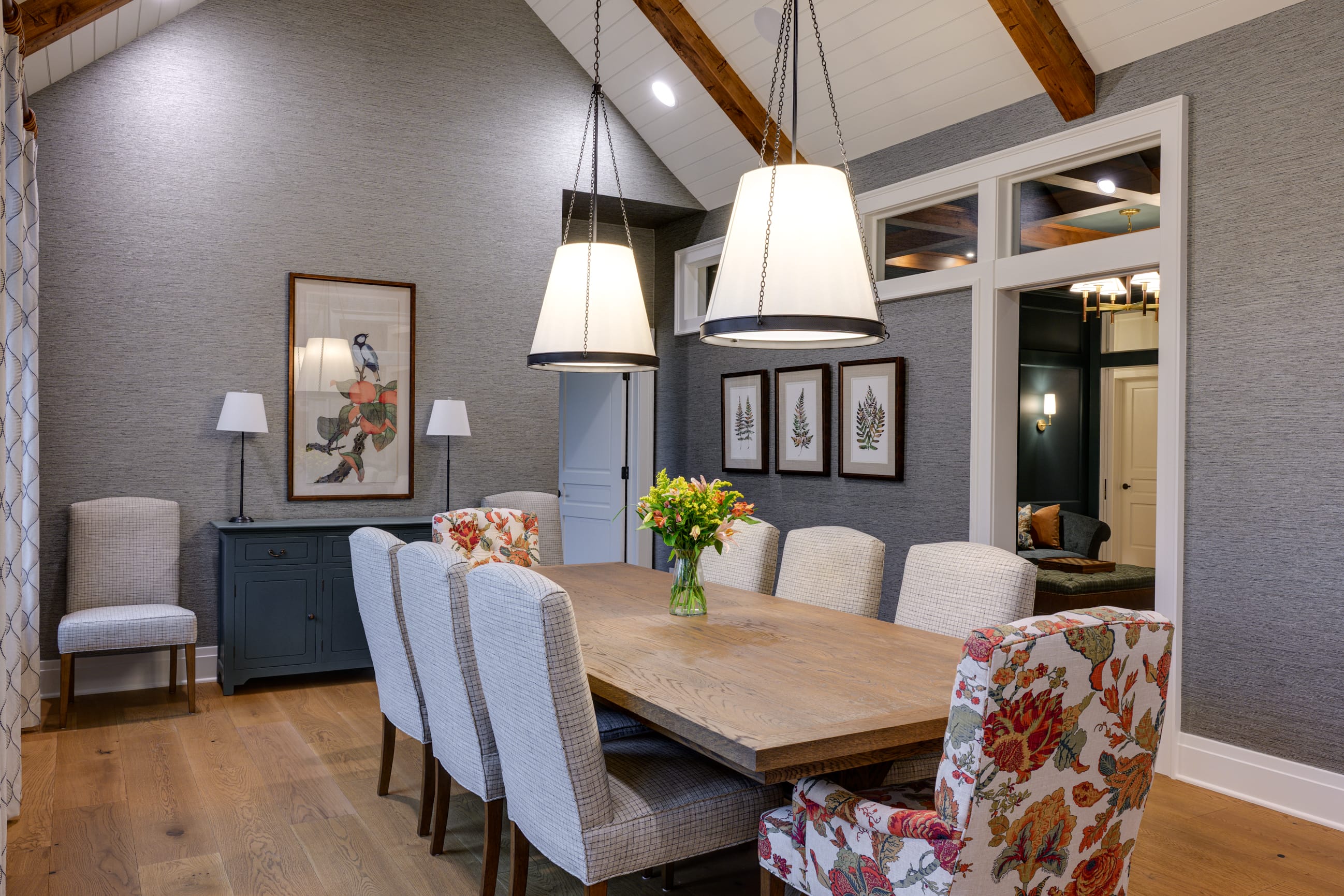 Bright dining area featuring a wooden table, floral chairs, and pendant lights, with artwork and greenery accents.