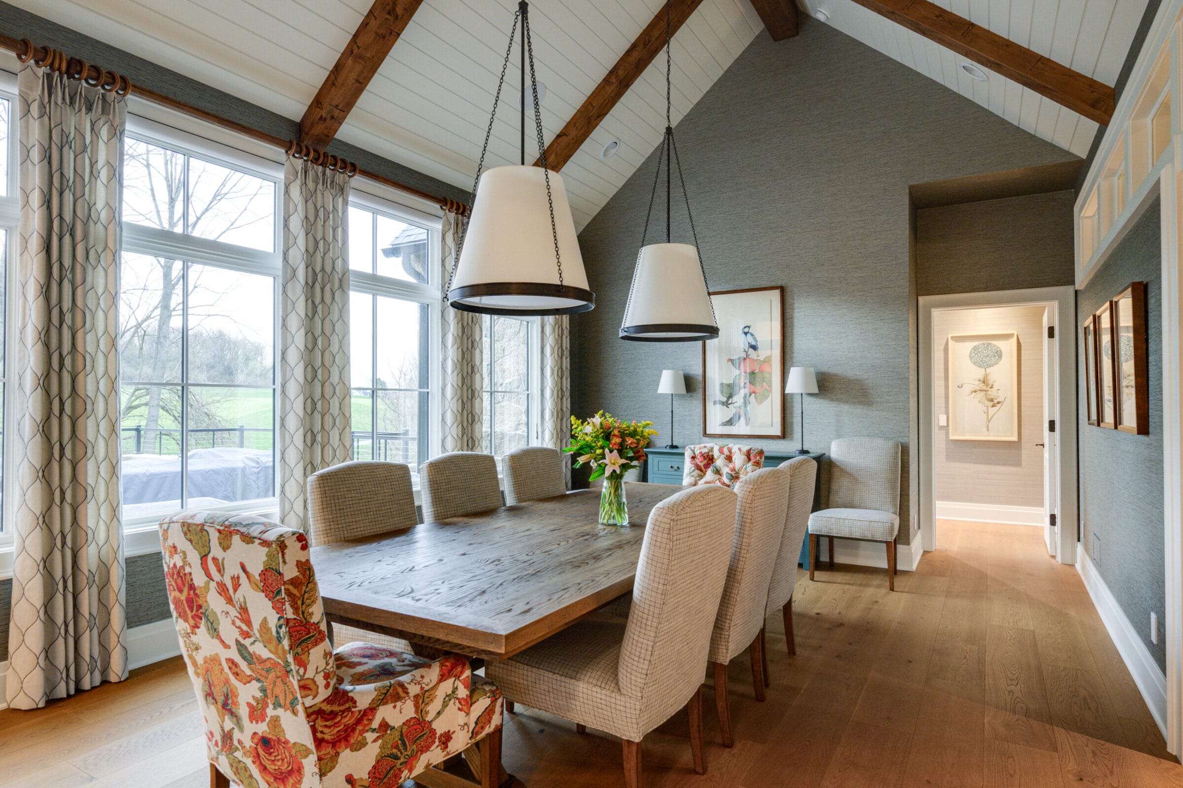 Bright dining room with a wooden table, upholstered chairs, large windows, and floral decor, featuring pendant lights above.