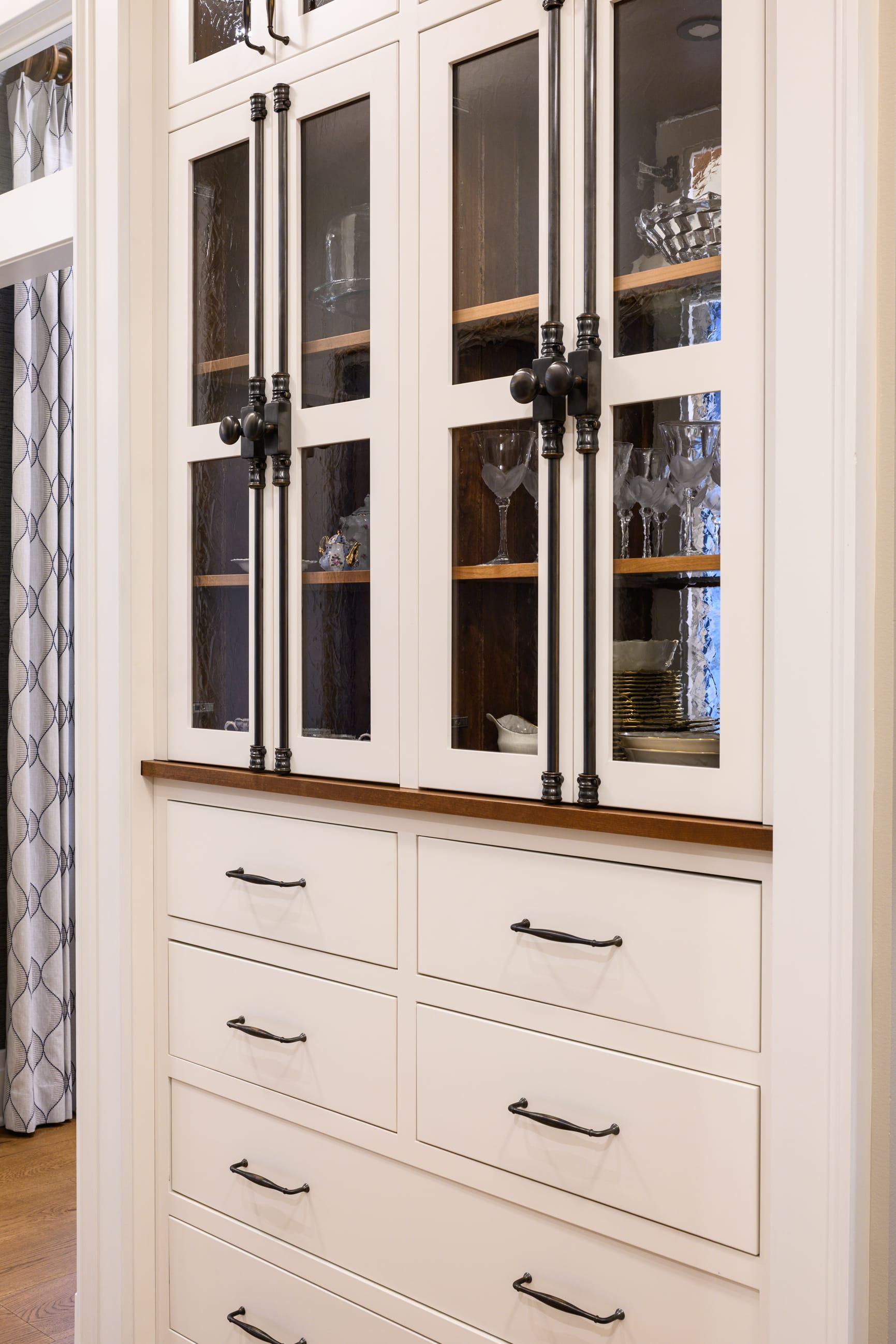 Elegant white cabinetry with glass doors displays glassware, complemented by a wooden countertop and sleek black handles.