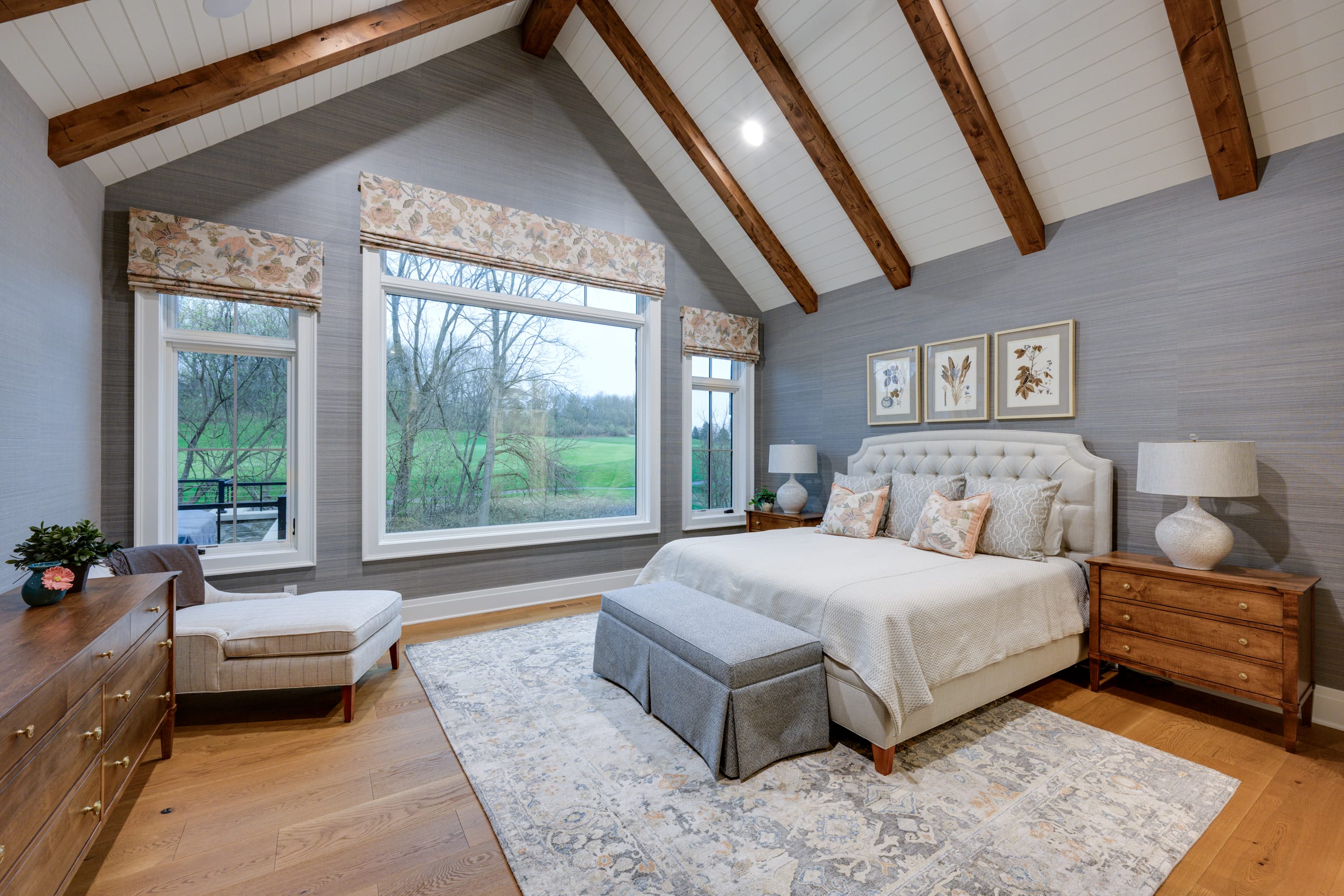 Cozy bedroom with vaulted wooden beams, large windows, a plush bed, and elegant decor overlooking a green landscape.