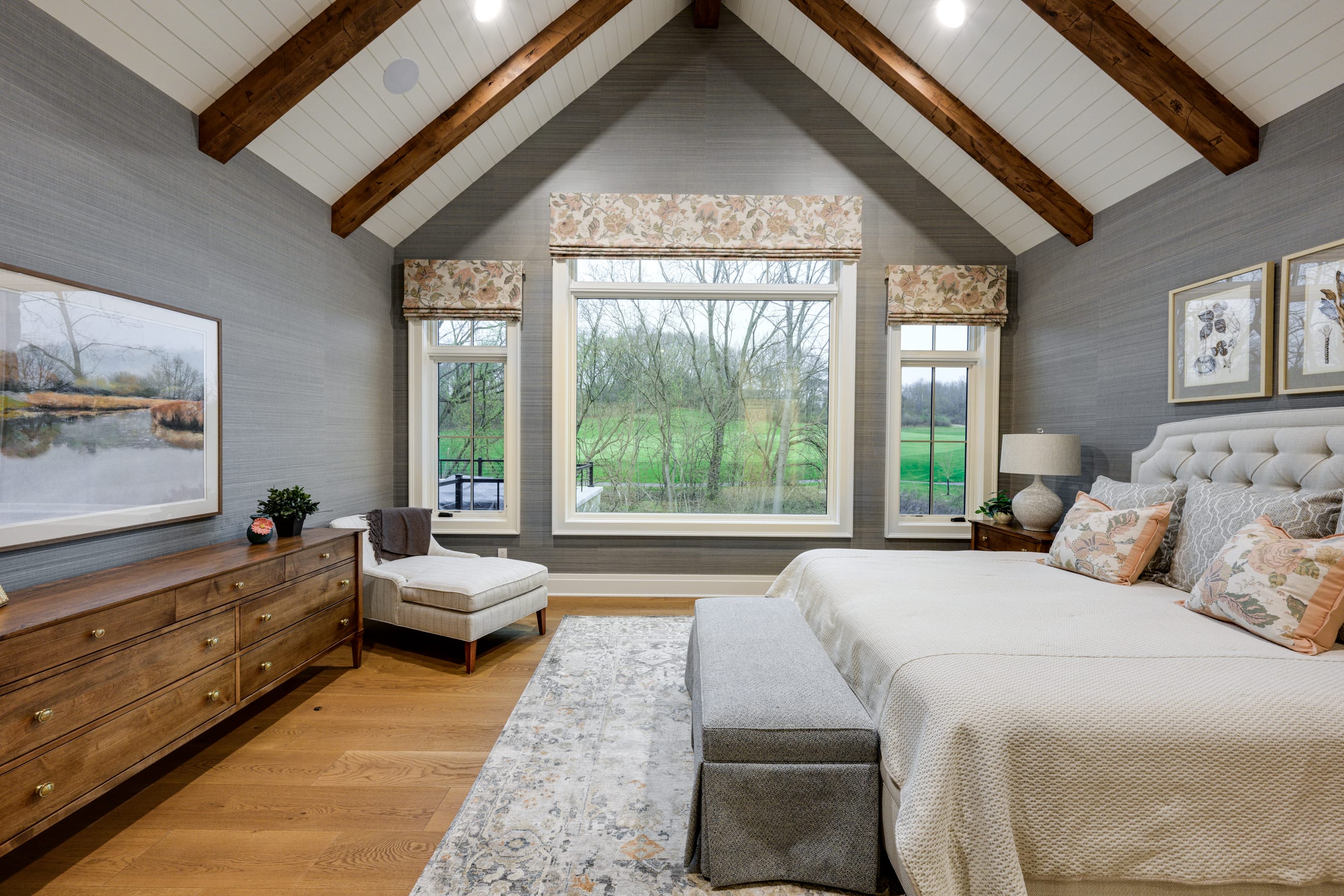 Cozy bedroom featuring a tufted bed, wooden dresser, large windows with floral shades, and a view of greenery outside.