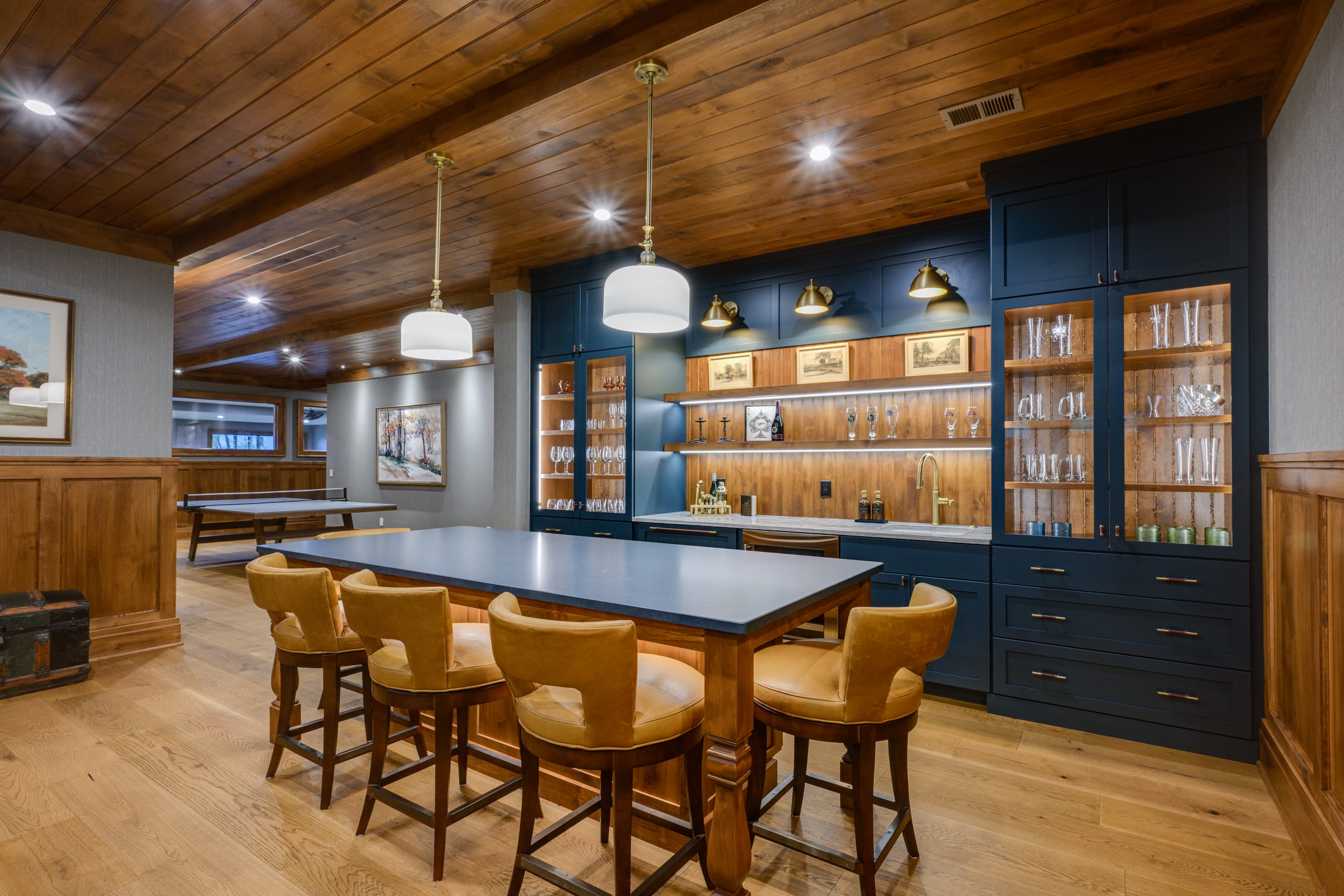 A warm, stylish home bar with a large wooden island, four tan leather bar stools, and two modern pendant lights overhead. The back wall features deep blue cabinetry with glass shelves displaying bottles and glassware, accented by brass fixtures and a brass faucet at the sink. The room has rich wood paneling, light hardwood floors, and a ping pong table and framed artwork visible in the background, creating a cozy, upscale recreation space.