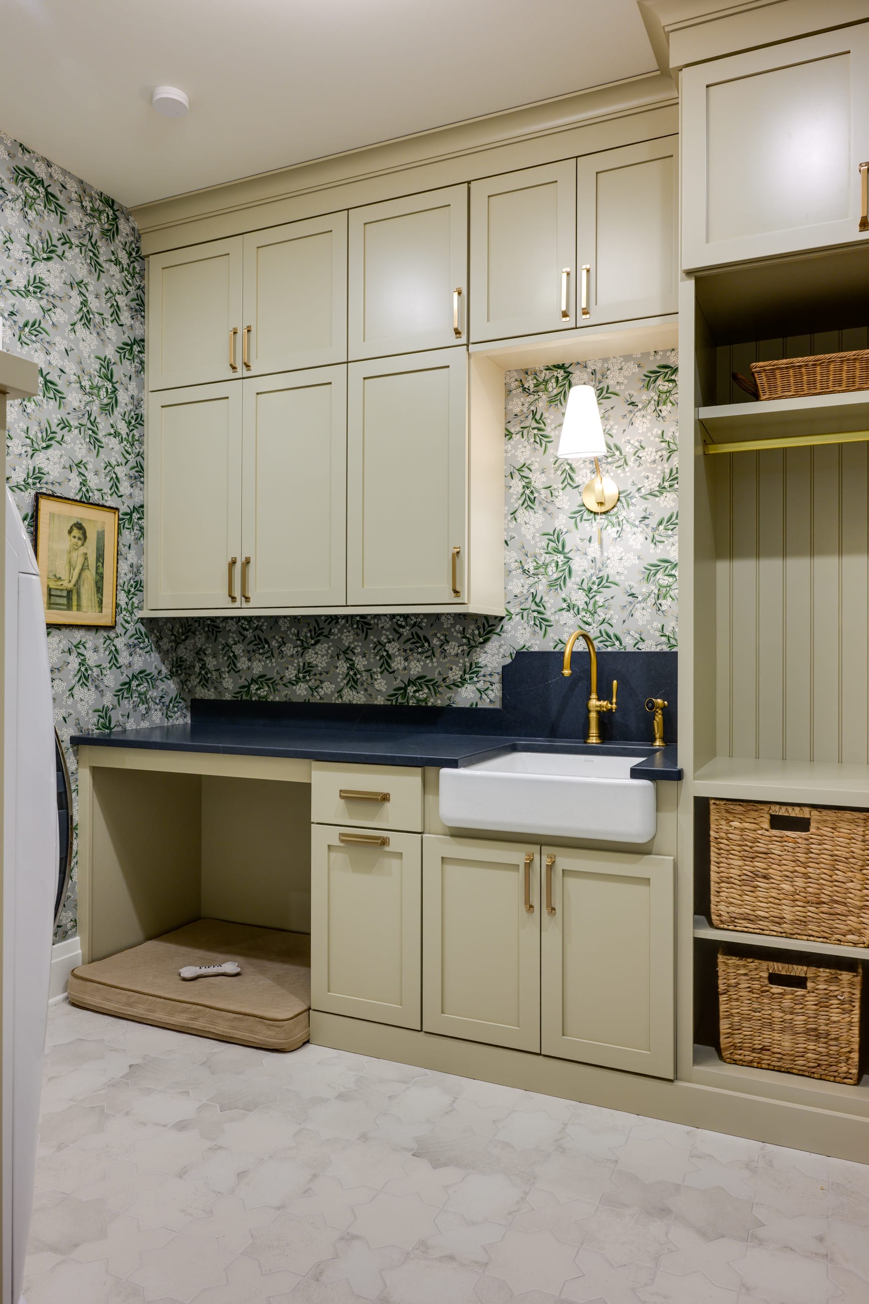 A stylish laundry room featuring pale green cabinets, a farmhouse sink, gold fixtures, and floral wallpaper.
