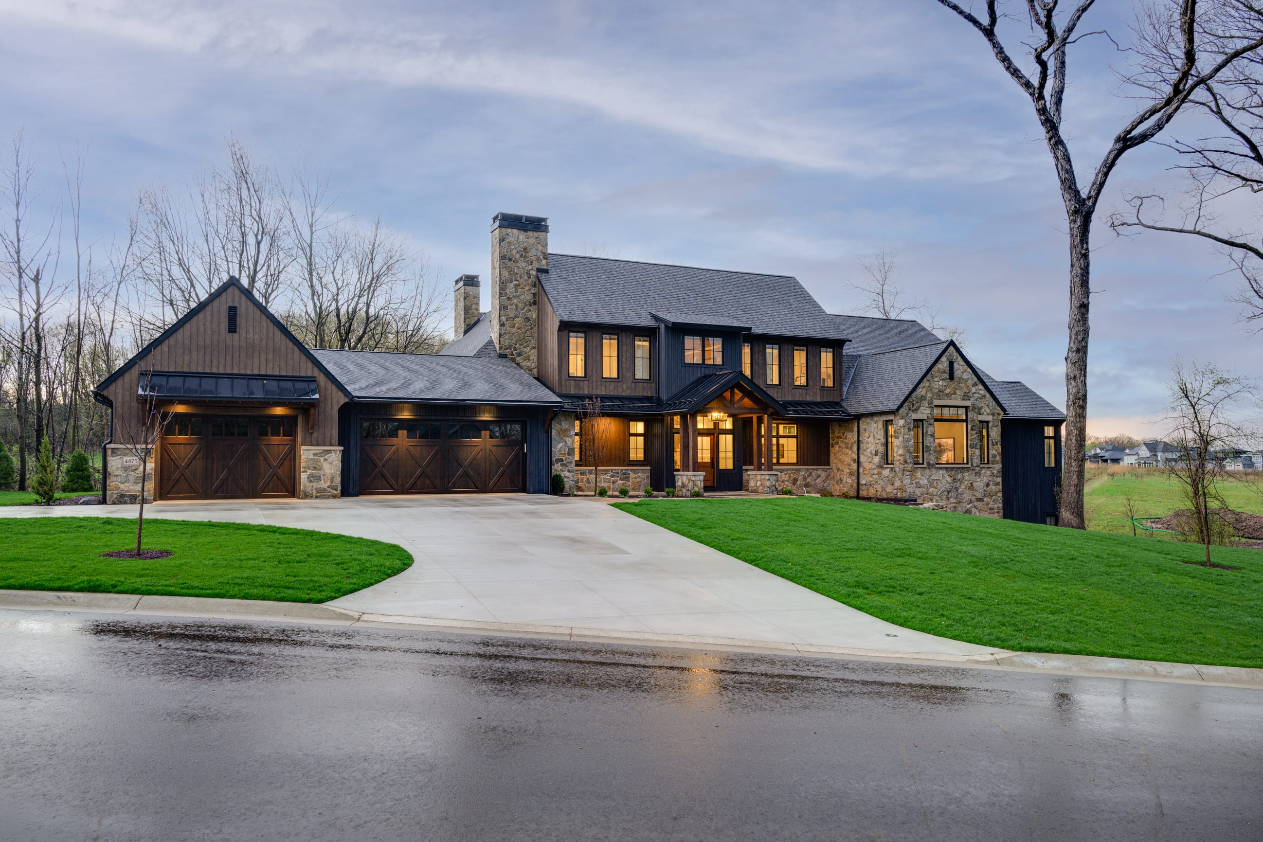 Modern stone and wood house with large windows, set on a green lawn, featuring a circular driveway and a tree nearby.