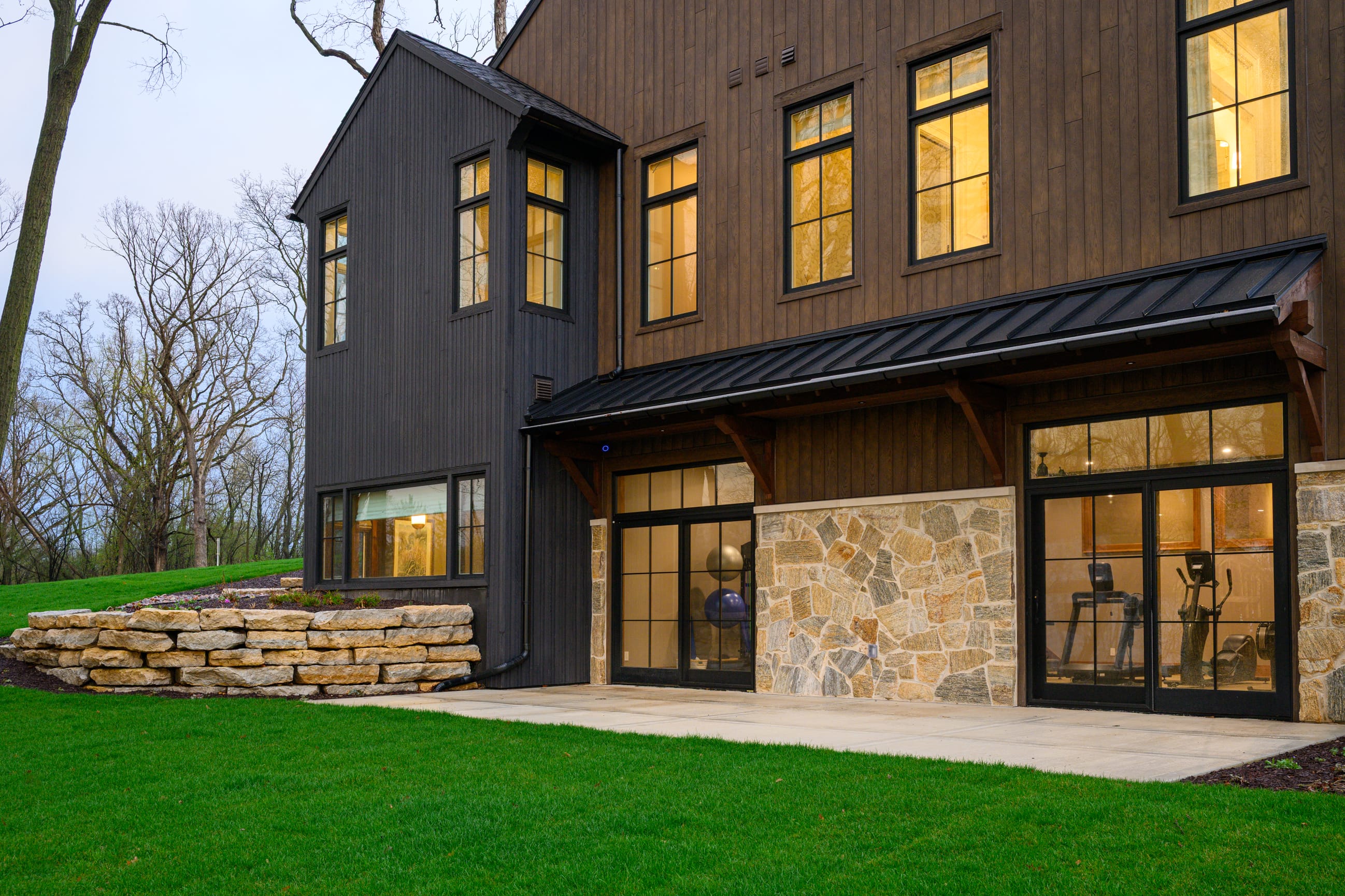 Modern two-story home with dark wood siding, large windows, and a stone foundation, set against a green lawn and trees.