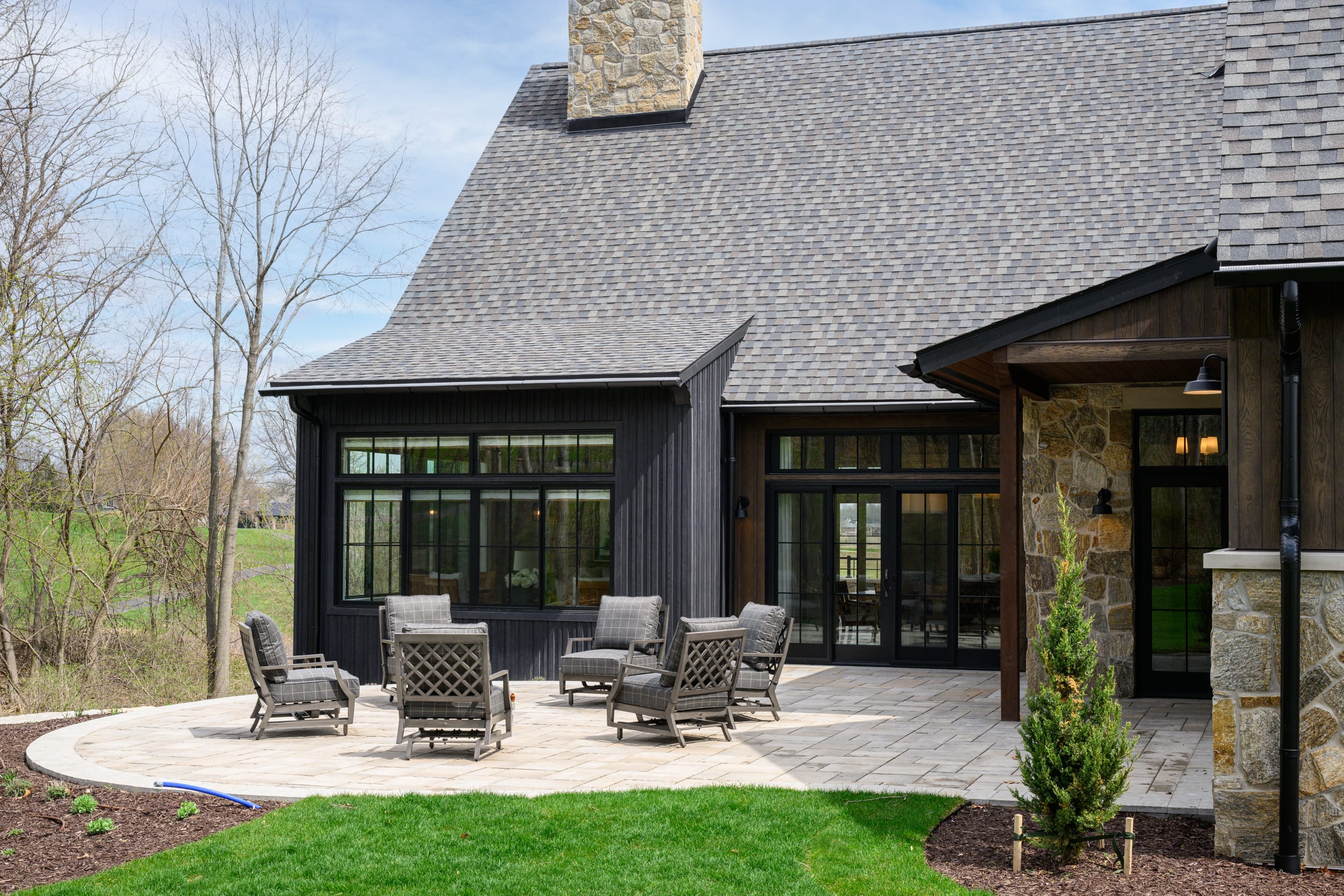 Modern patio with stone and wood accents, featuring gray lounge chairs and a manicured lawn under a clear blue sky.