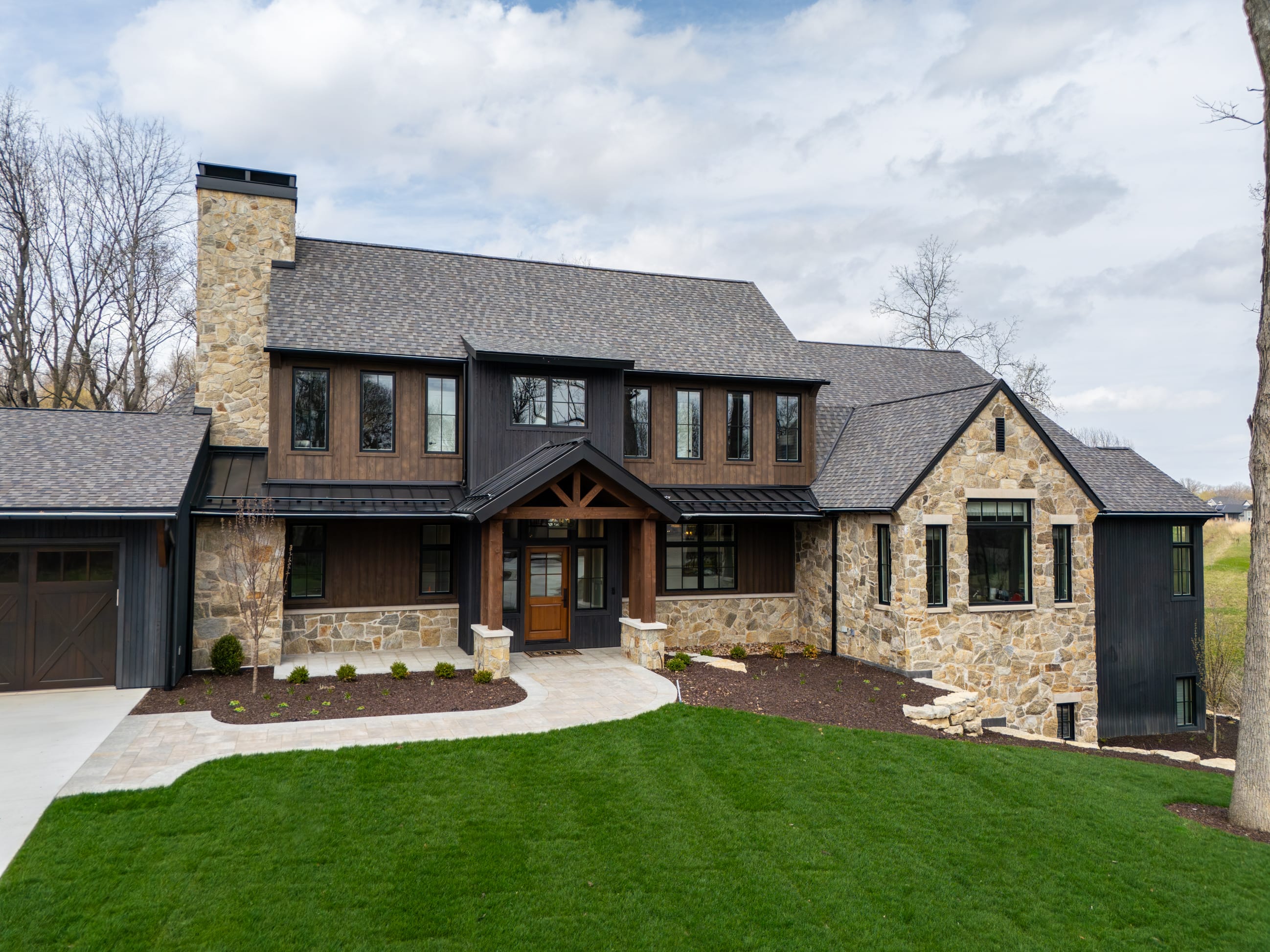 Modern stone and wood house with a sloped roof, large windows, and a manicured lawn under a cloudy sky.