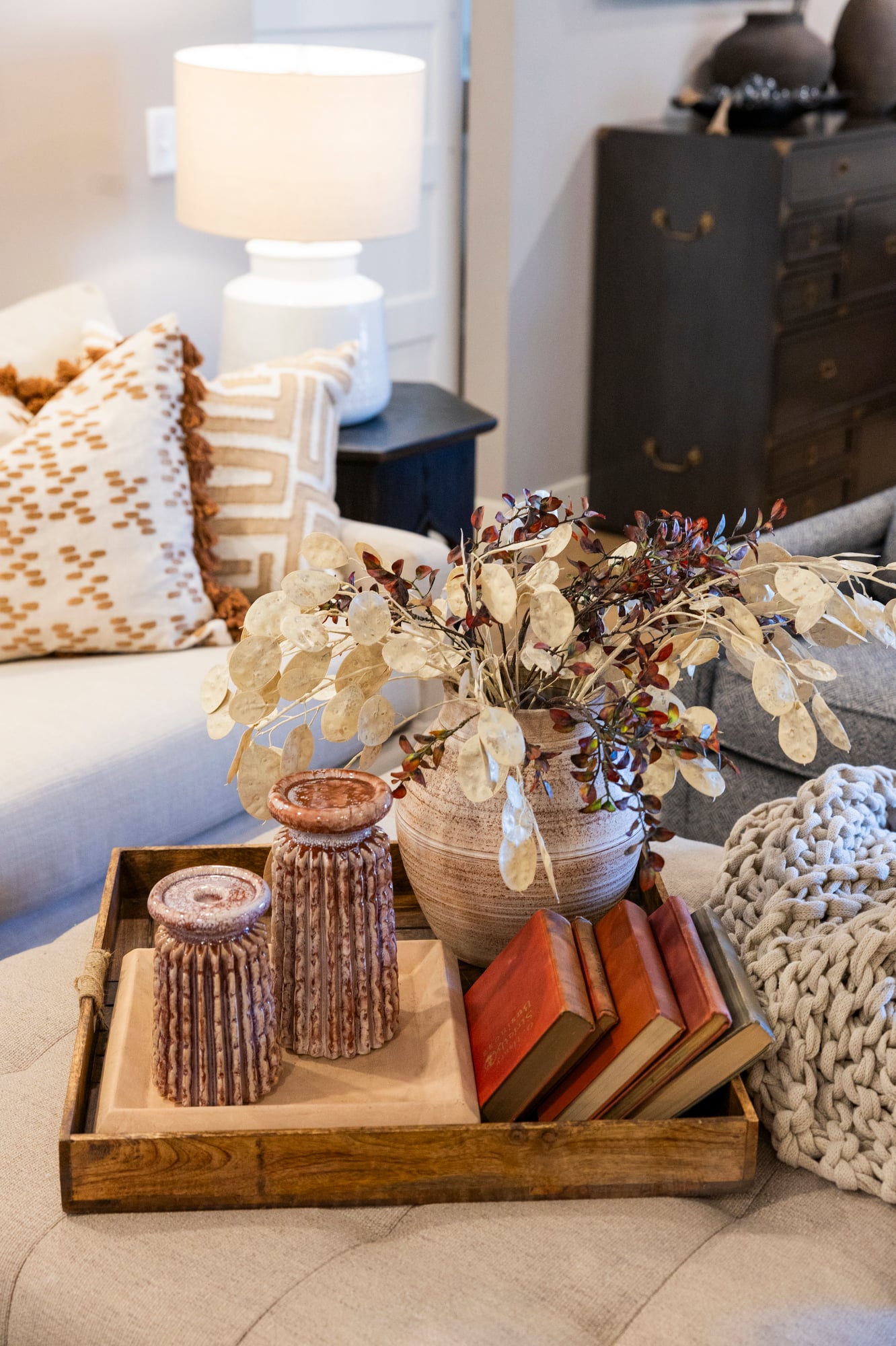 Wooden tray with decorative items: vase with dried foliage, two carved candle holders, and stacked books.