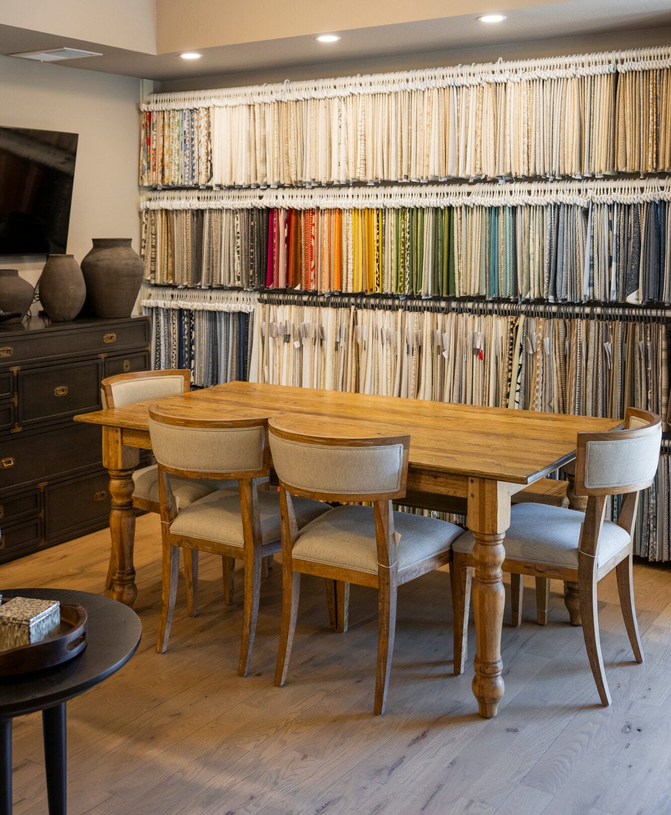 Wooden dining table with upholstered chairs in front of a wall of fabric swatches.
