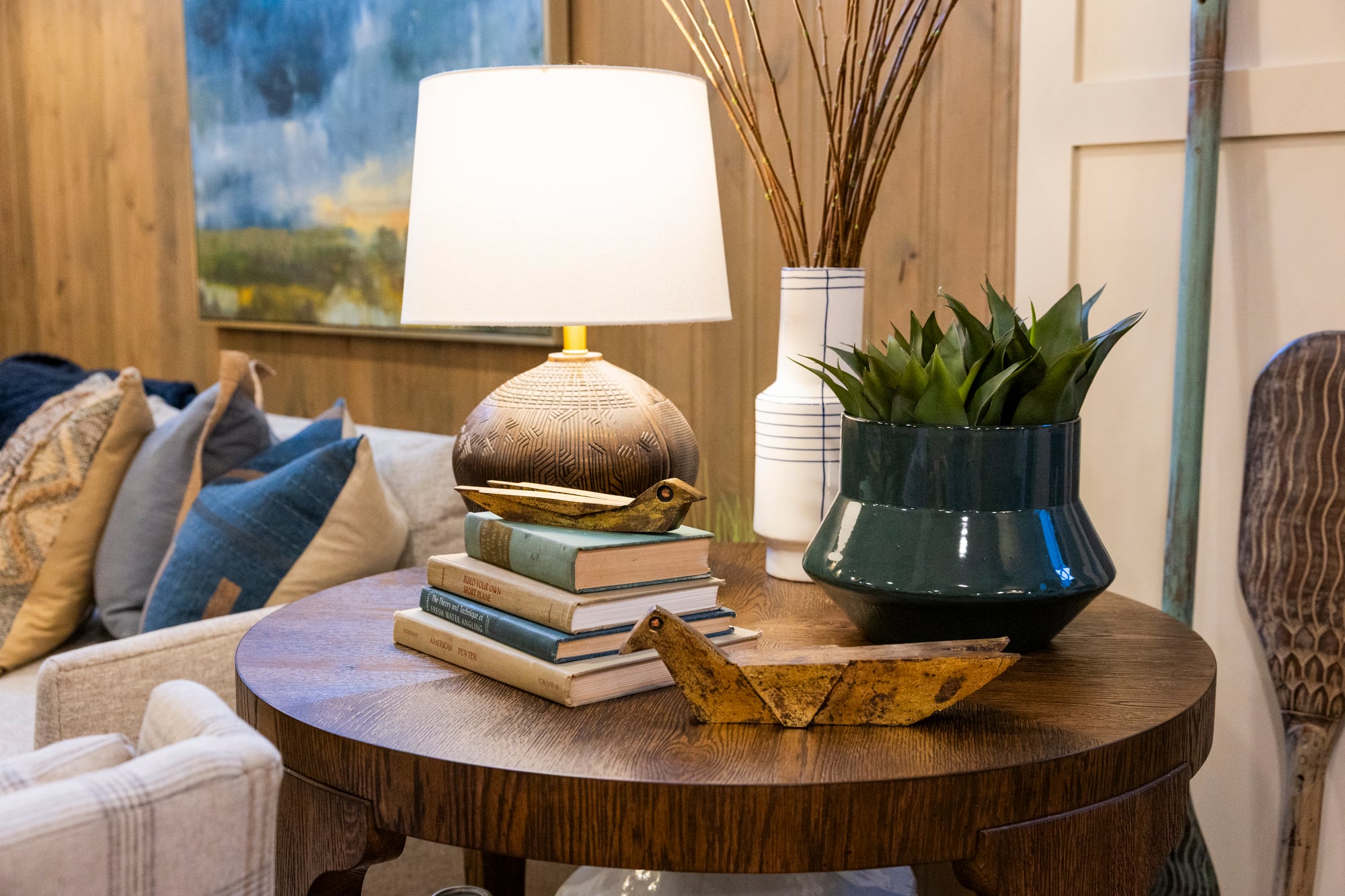 Round wooden table with lamp, books, plant, and decorative birds.