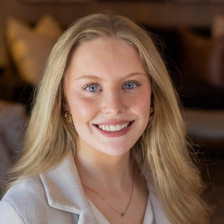 Blonde woman with blue eyes smiling, wearing a light jacket and gold earrings.
