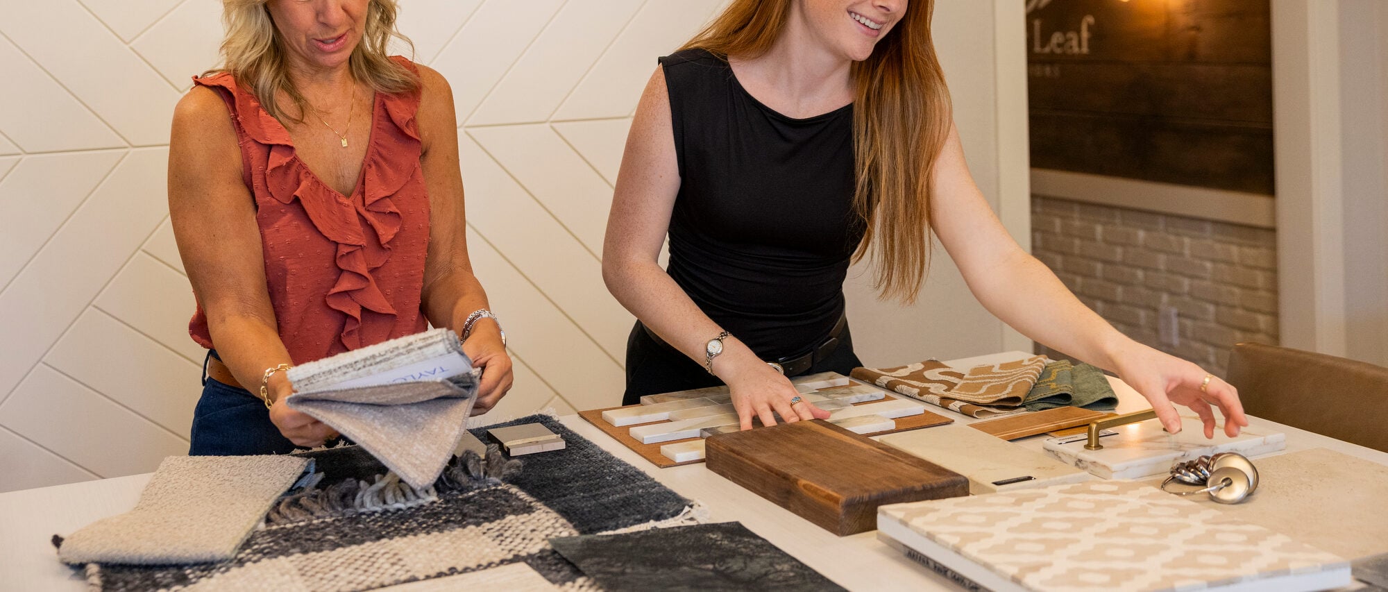 Two women review fabric and tile samples on a table.