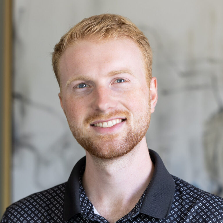 Man with red hair and blue eyes smiling, wearing a black patterned shirt.