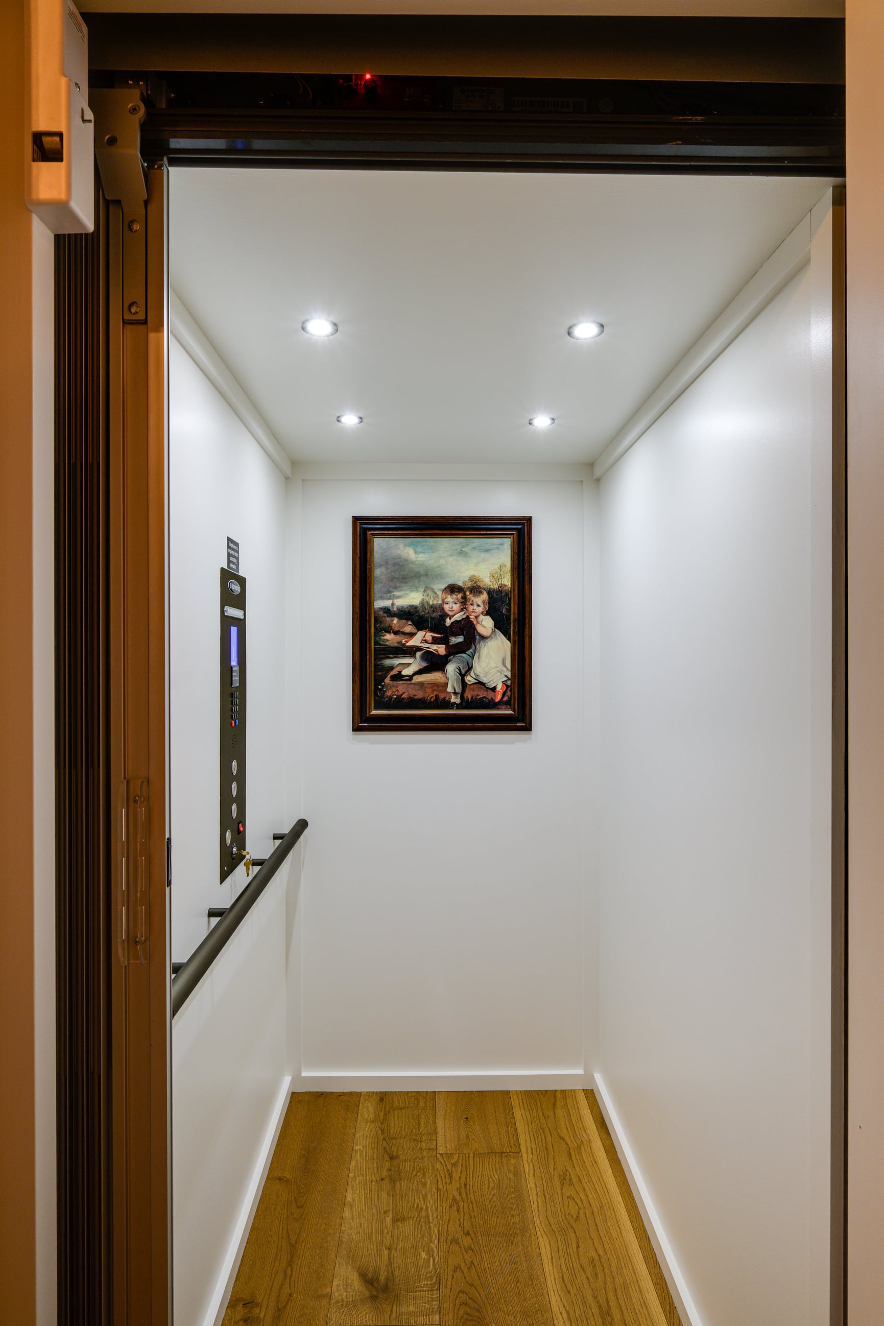 The inside of a small home elevator with wood floors and white walls. There is a painting of two young children hanging on the wall across from the entrance.