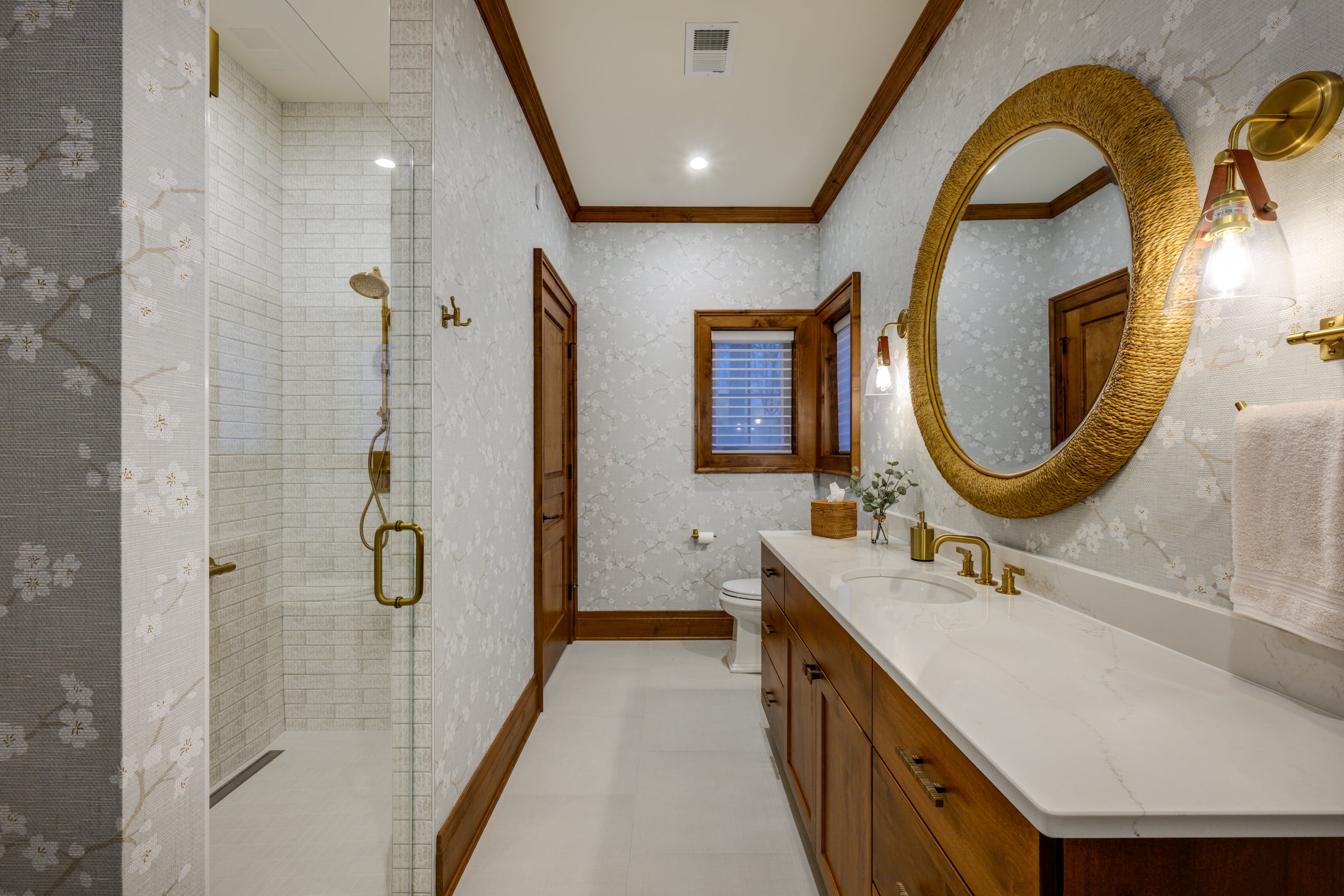 Bathroom with floral wallpaper, glass shower, and wooden vanity with gold fixtures.