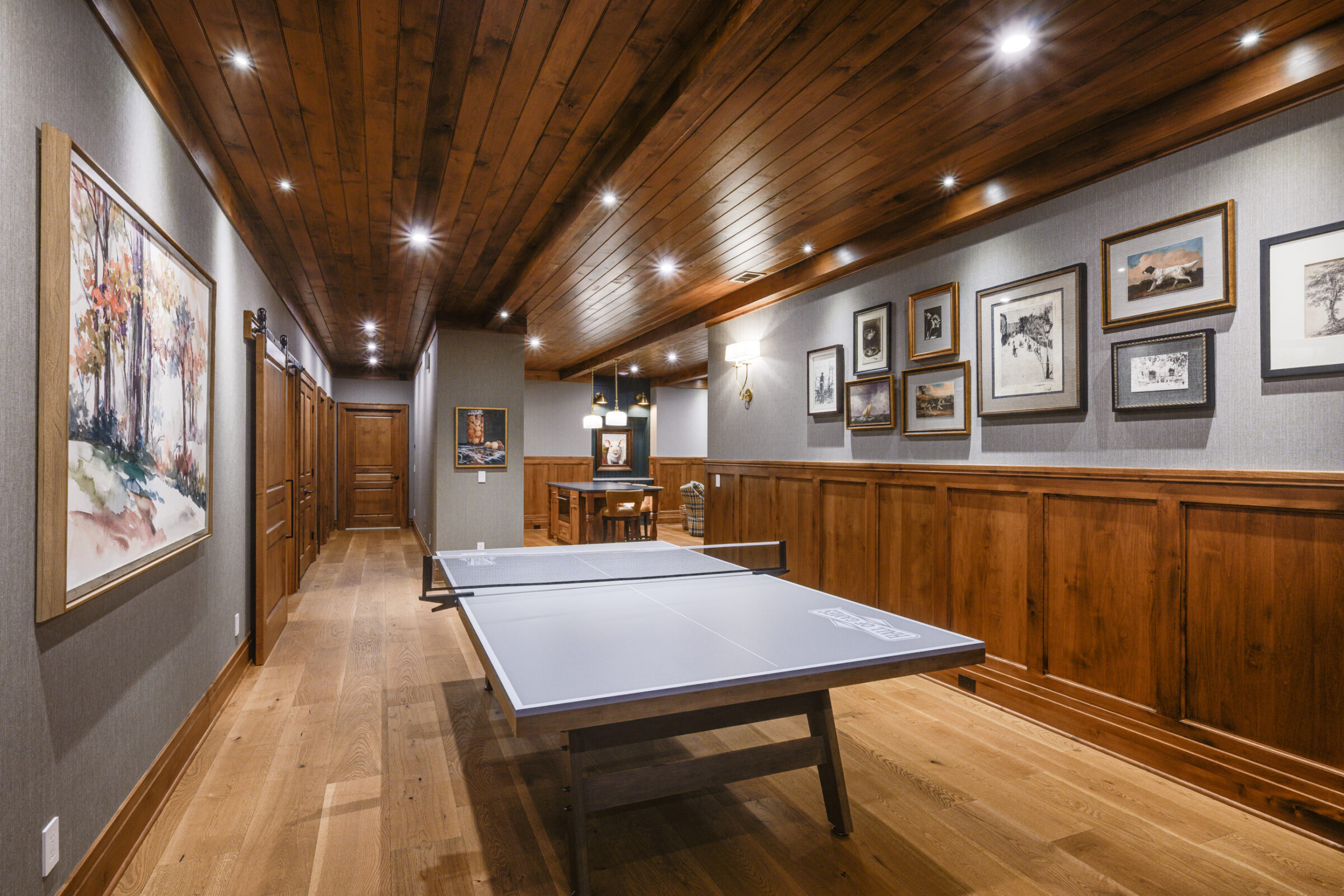 Ping pong table in a wood-paneled room with artwork and a long hallway.
