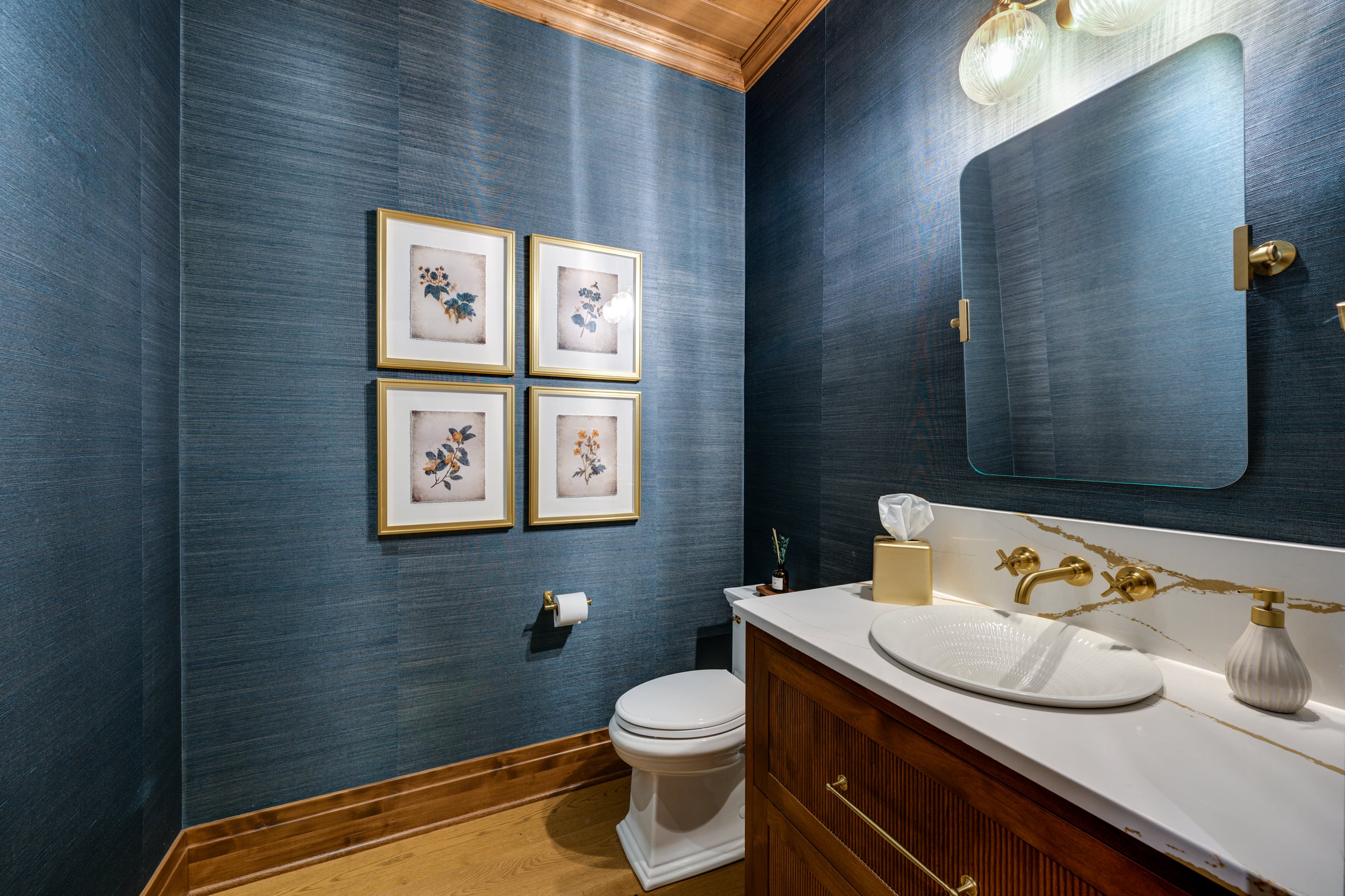 Powder room with blue textured wallpaper, gold-framed botanical prints, and a wooden vanity with a white sink.