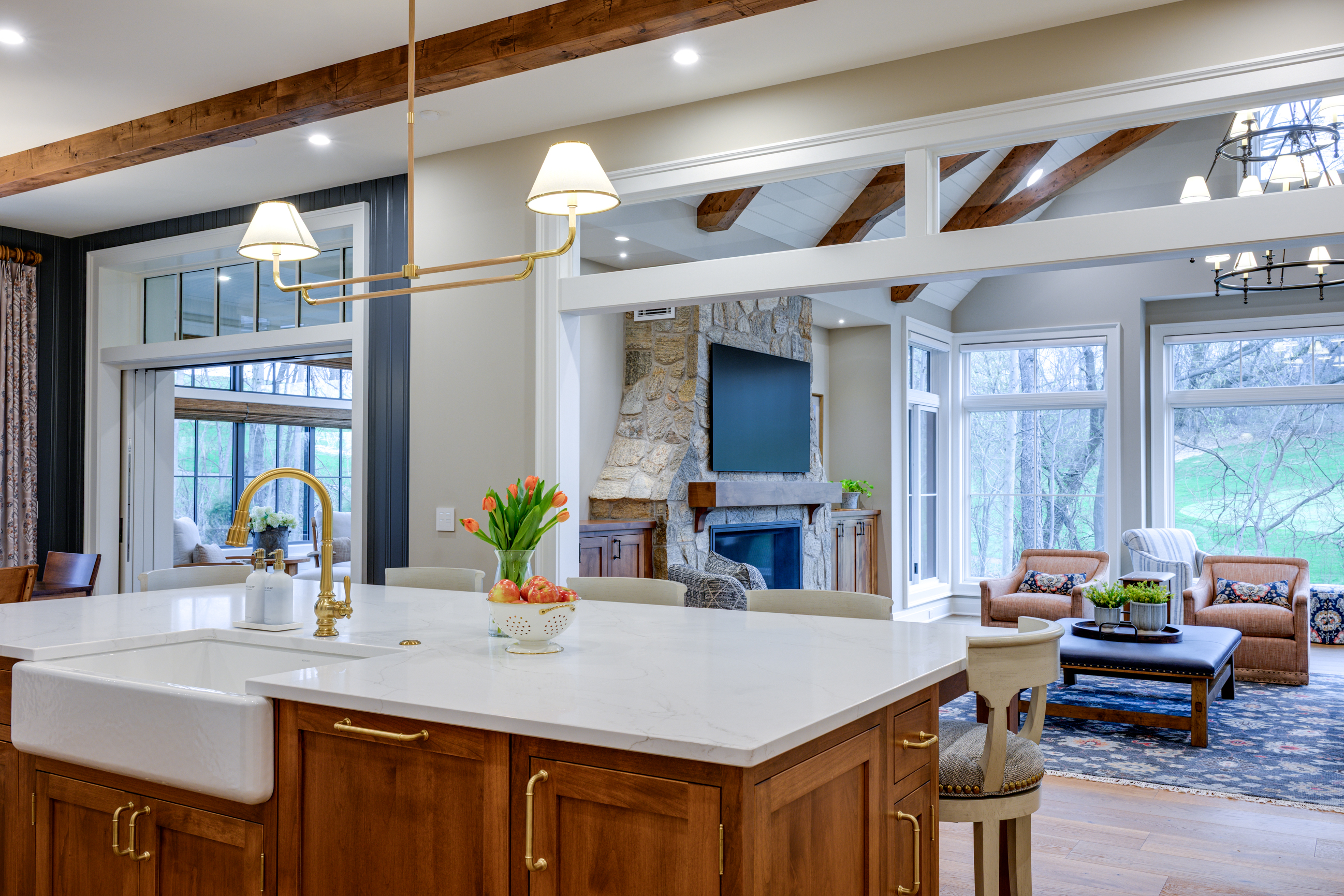 Kitchen island with white sink and gold faucet, facing a living room with stone fireplace and large windows.
