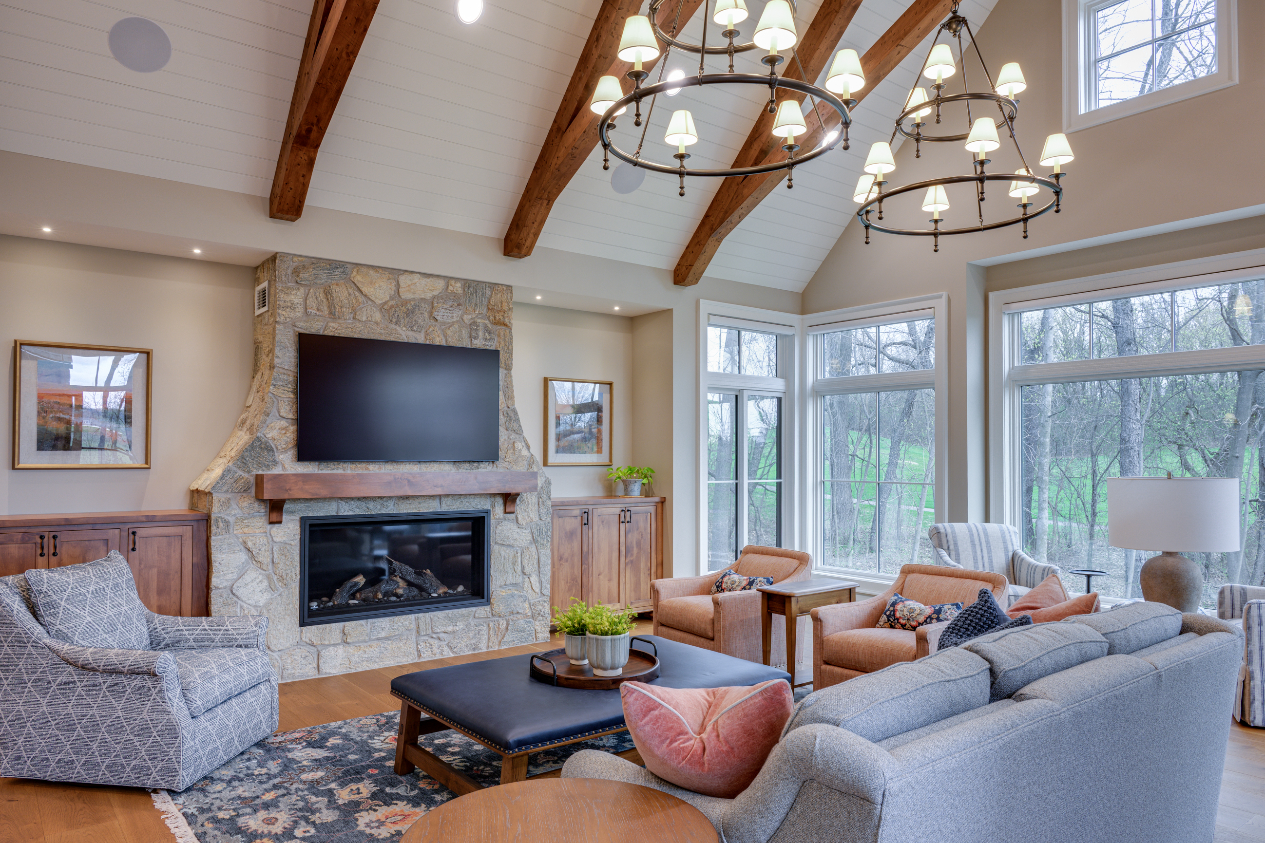 Living room with stone fireplace, large TV, vaulted beamed ceiling, and large windows overlooking trees.