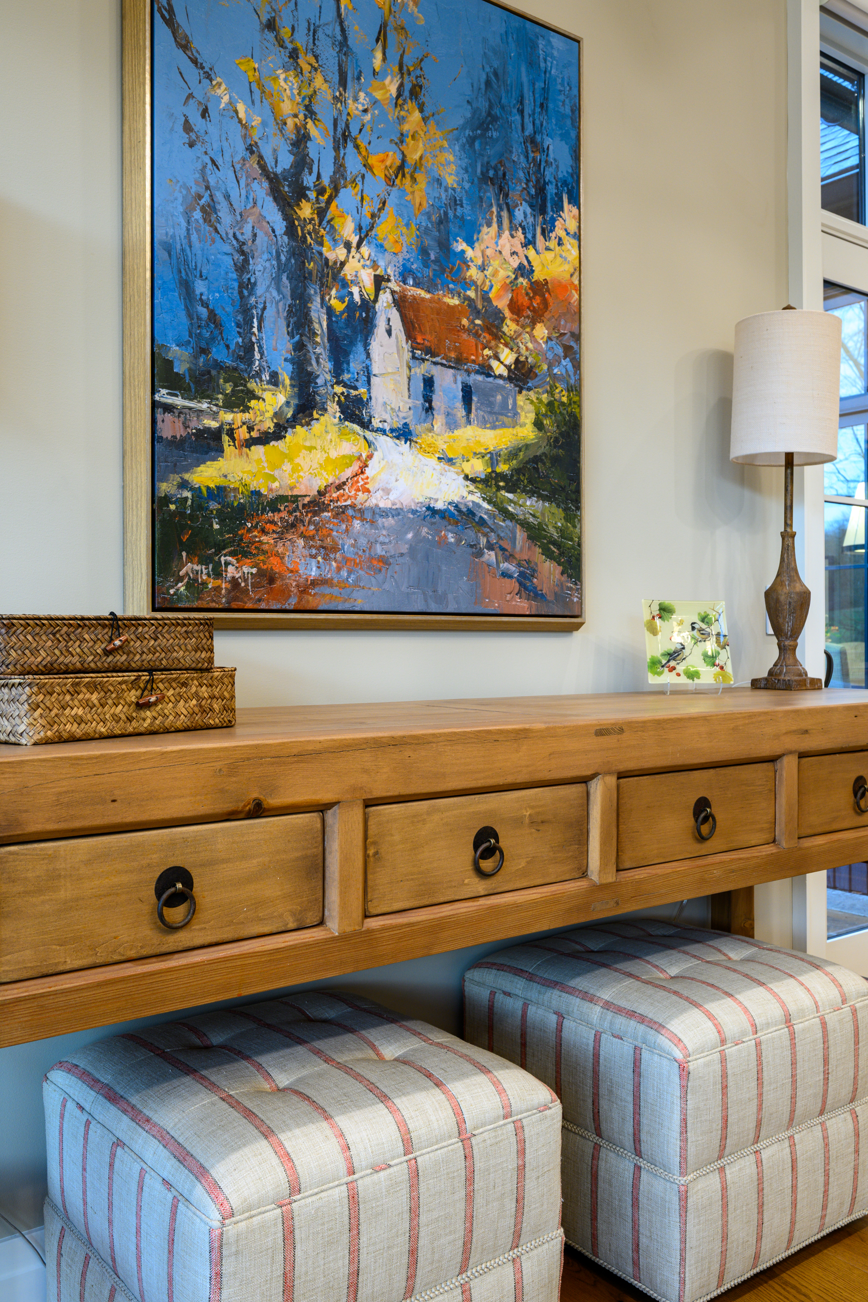 Painting of a house and trees above a wooden console table with striped ottomans.