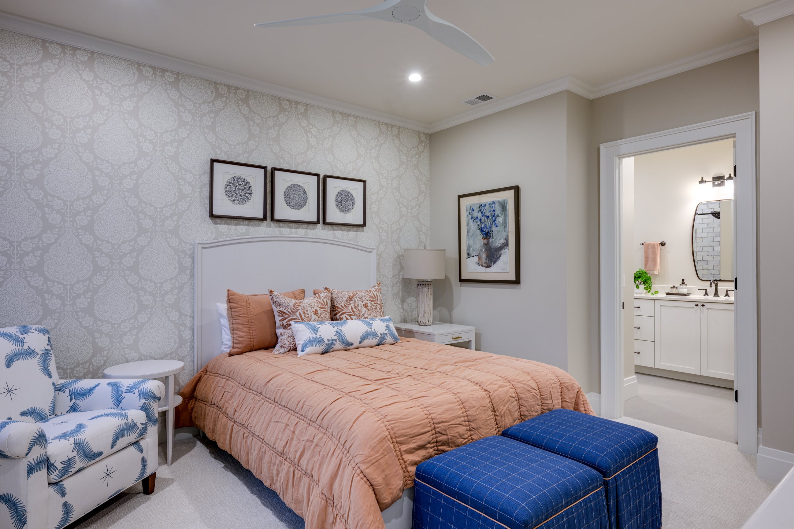 Bedroom with peach quilt, blue ottomans, patterned chair, and doorway to bathroom.