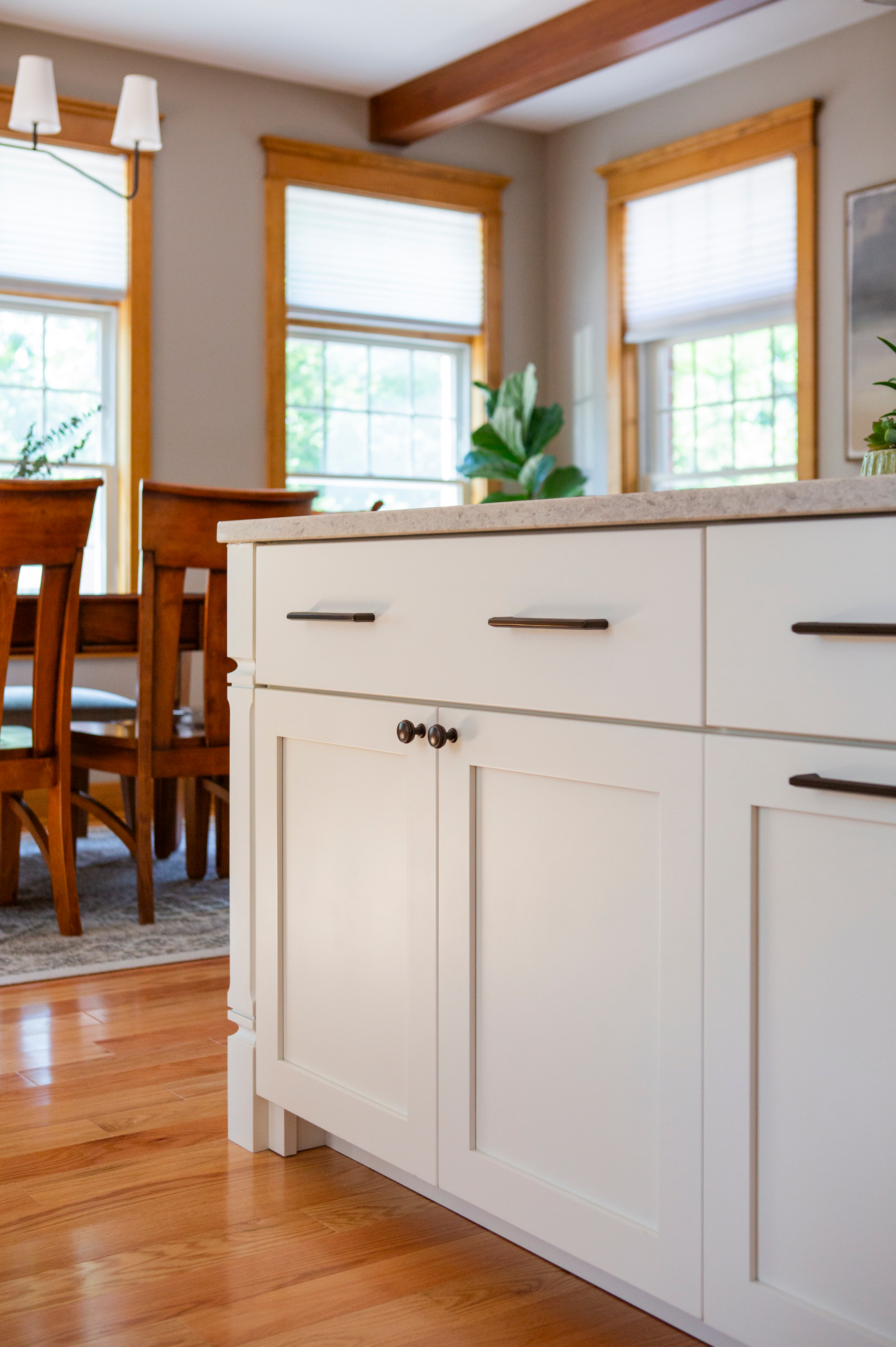 White kitchen island with dark hardware, wooden dining chairs, and windows.