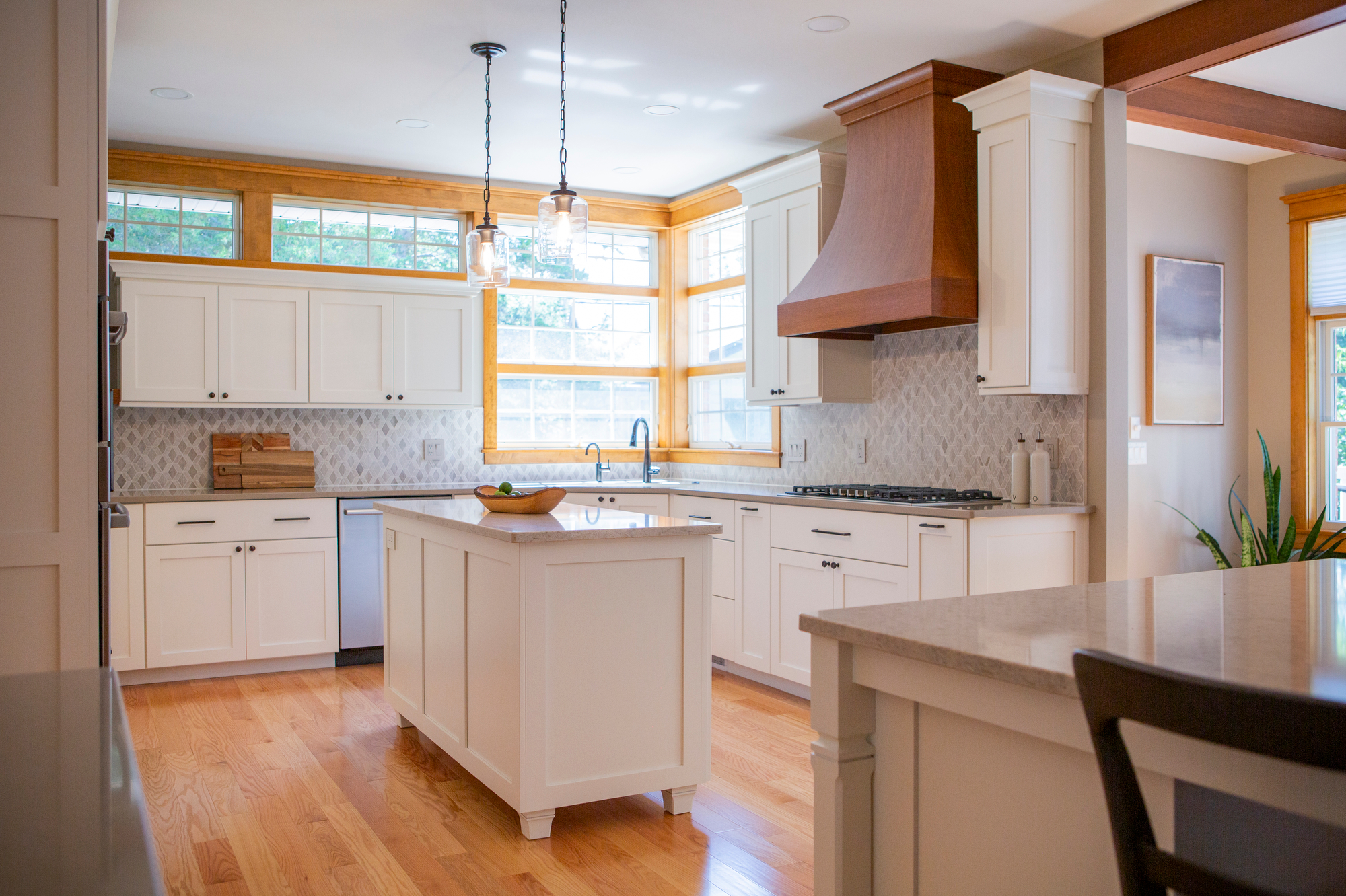 Modern white kitchen with island, wood range hood, and large windows.