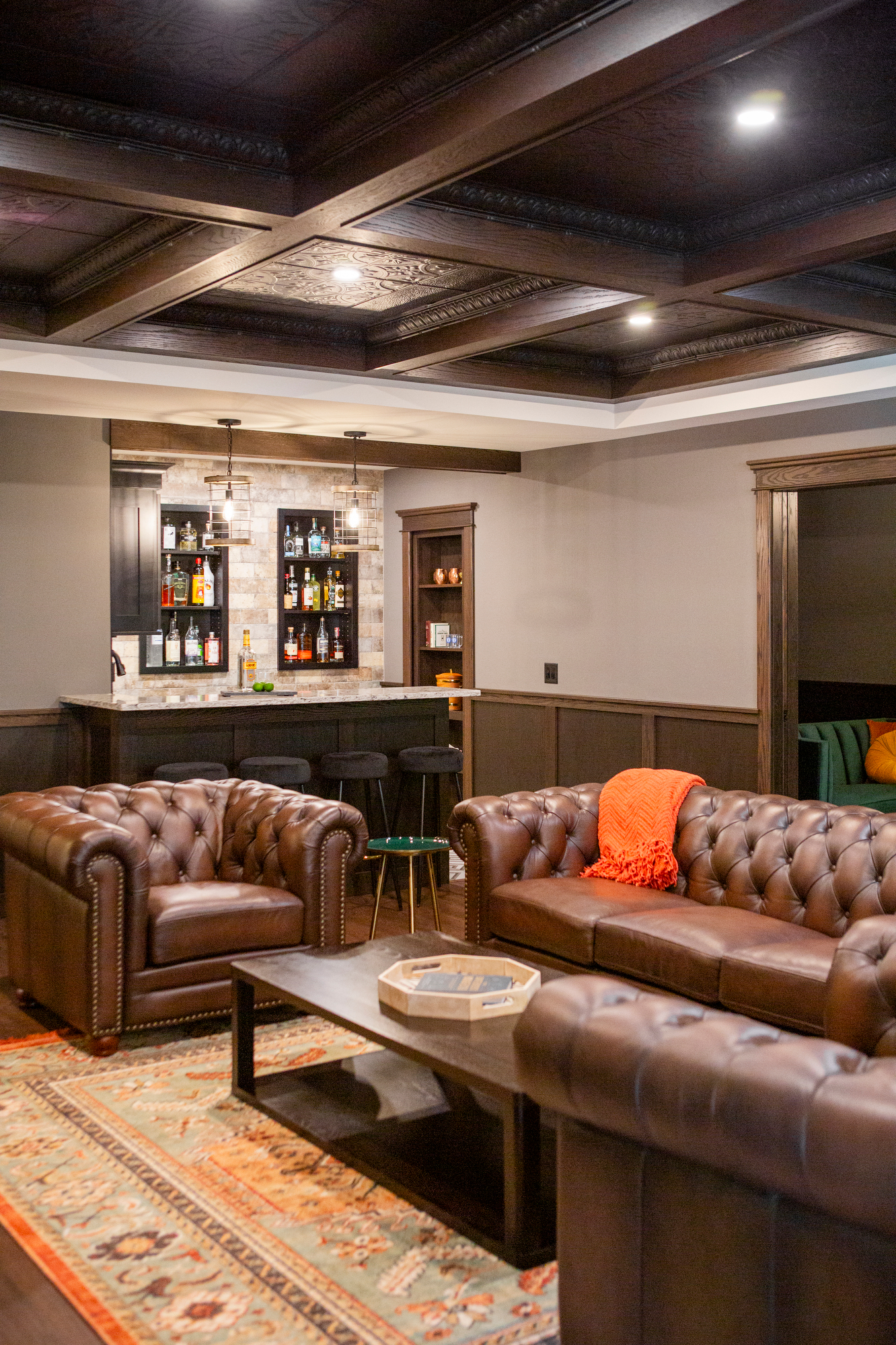 Leather sofas and armchairs in a room with a bar, coffered ceiling, and patterned rug.