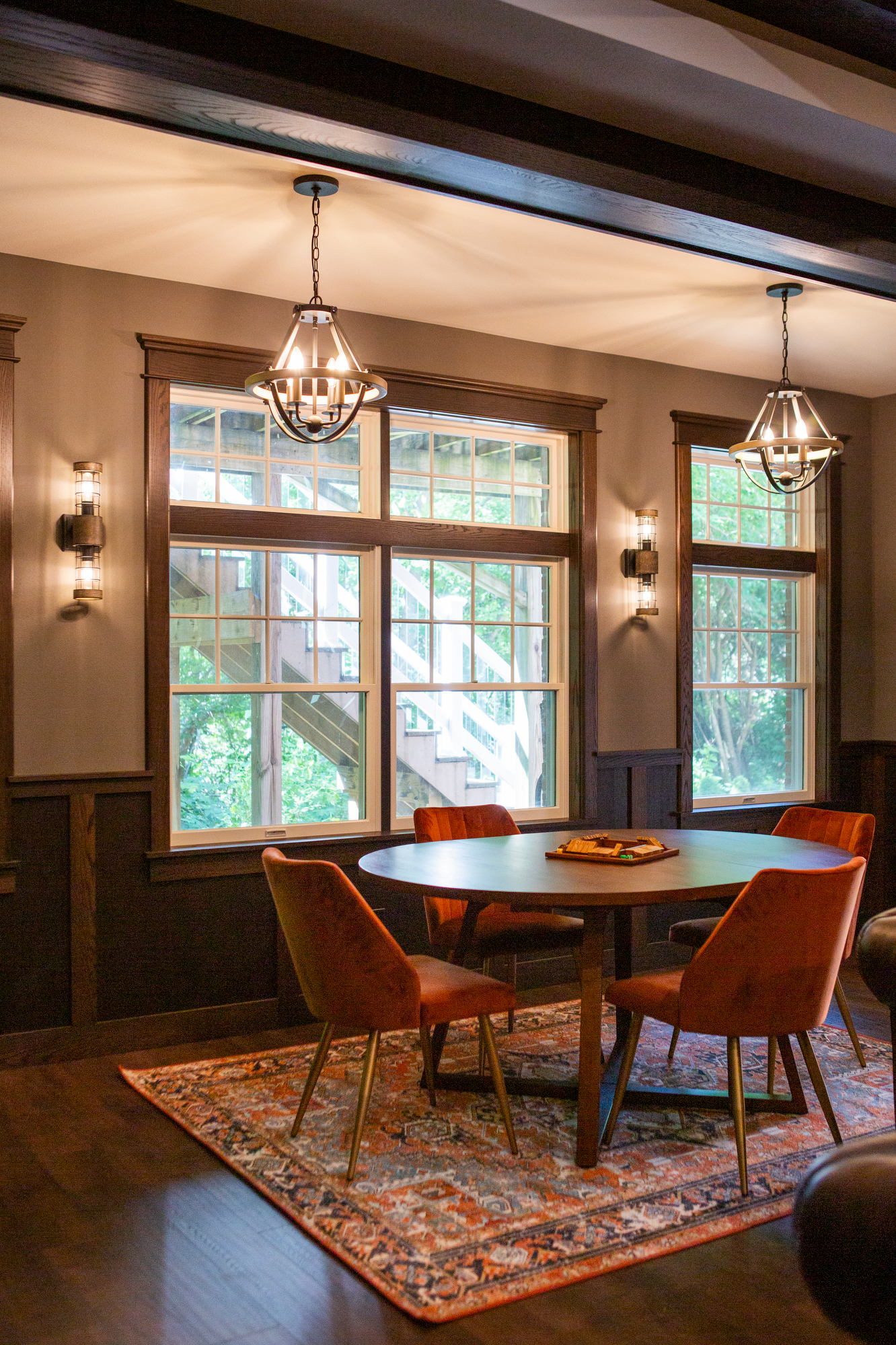 Round table with orange chairs on patterned rug, near large windows.