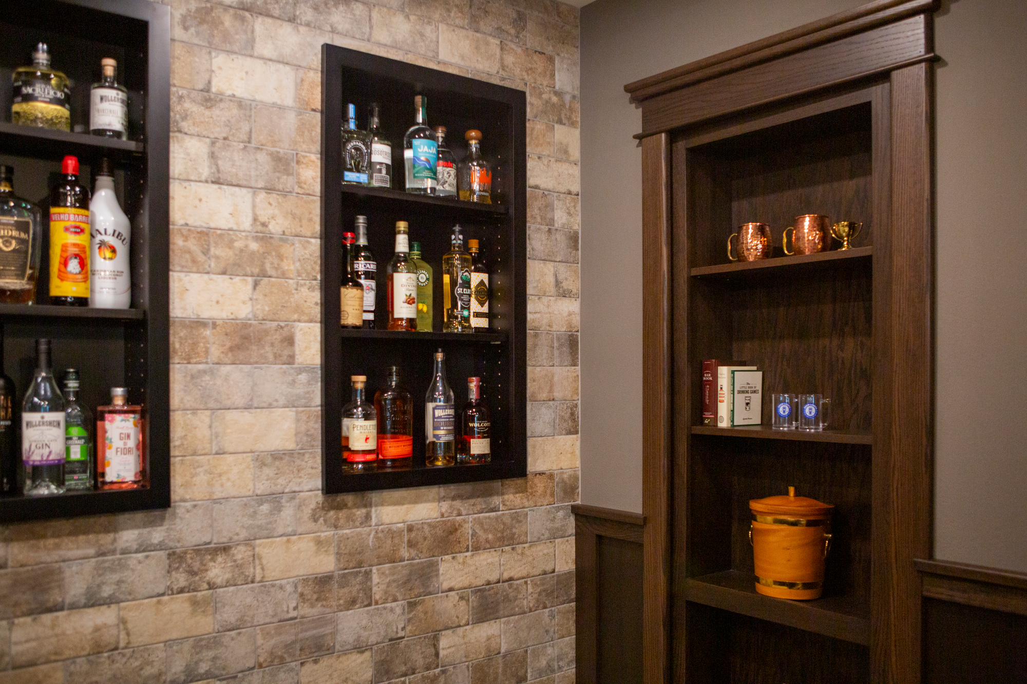 Shelves of liquor bottles on a brick wall next to a wooden bookshelf with mugs and glasses.