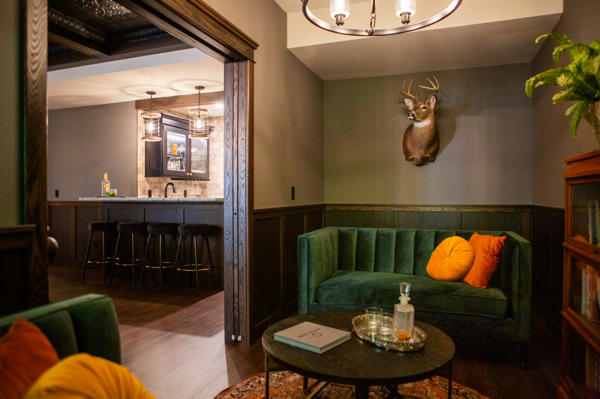 Green velvet sofa with orange pillows, deer head mount, and bar area.