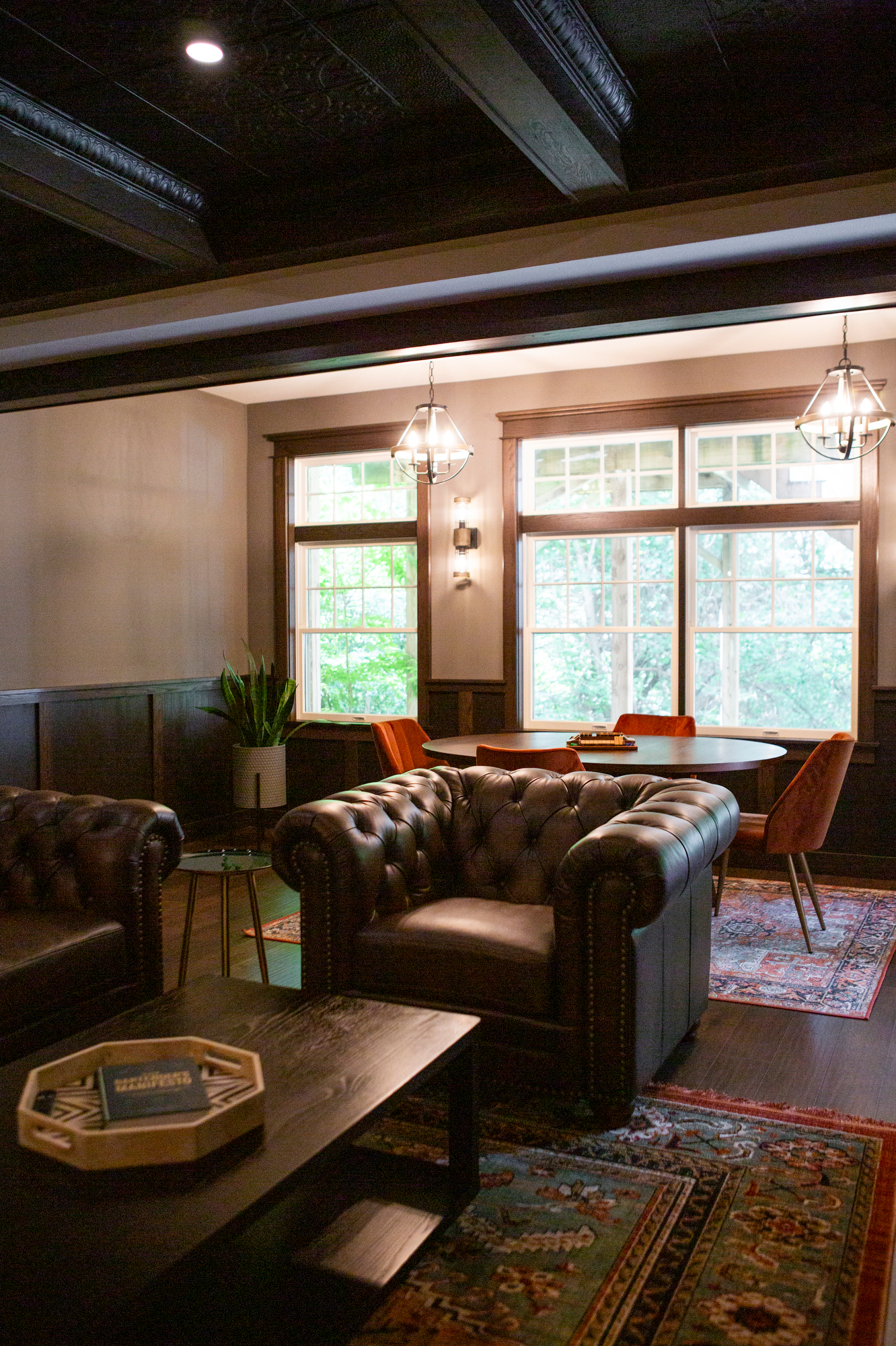 Chesterfield armchairs and a round table with orange chairs in a room with large windows.