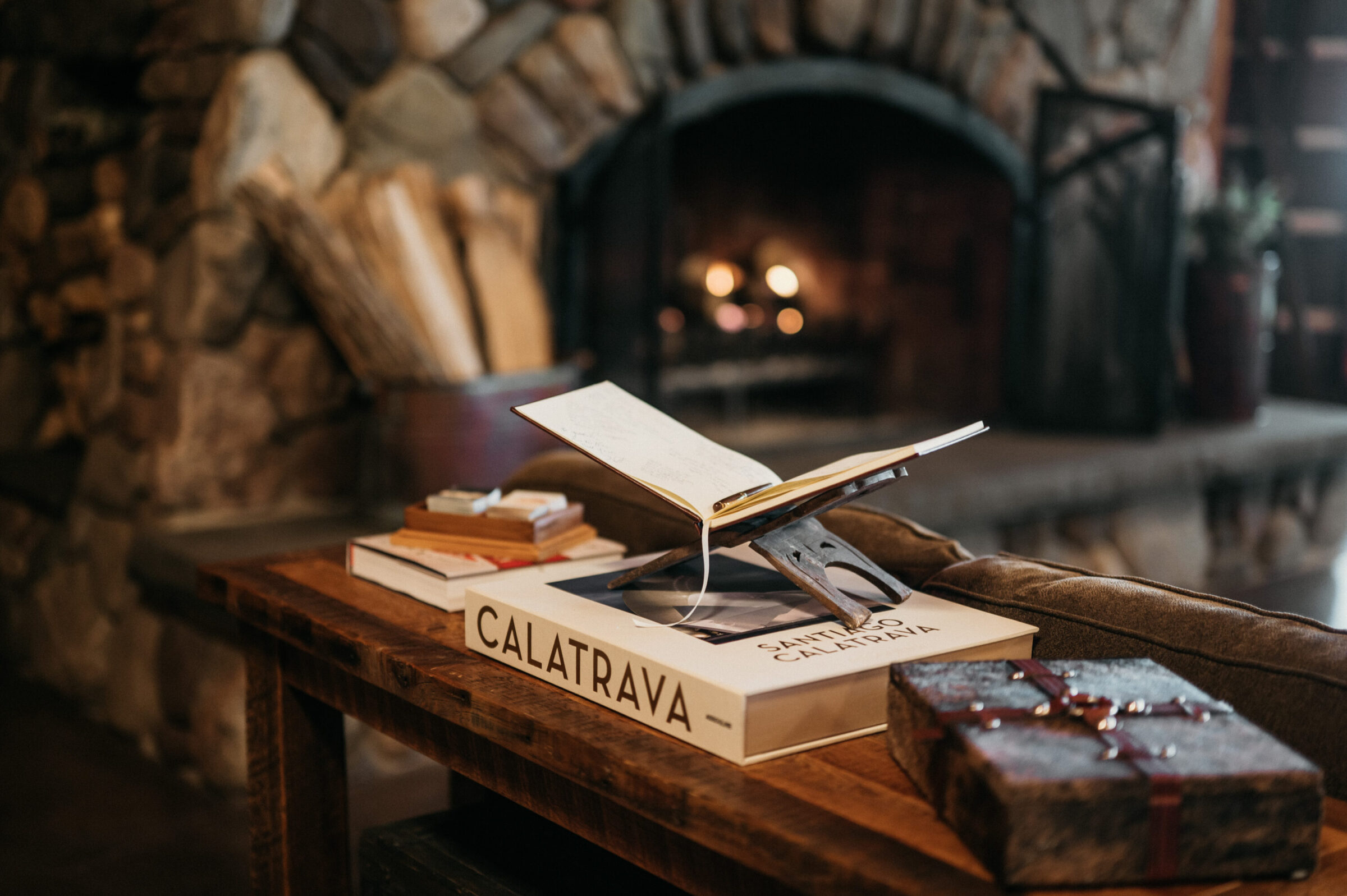 Open book on stand, books, and decorative box on wooden table in front of fireplace.