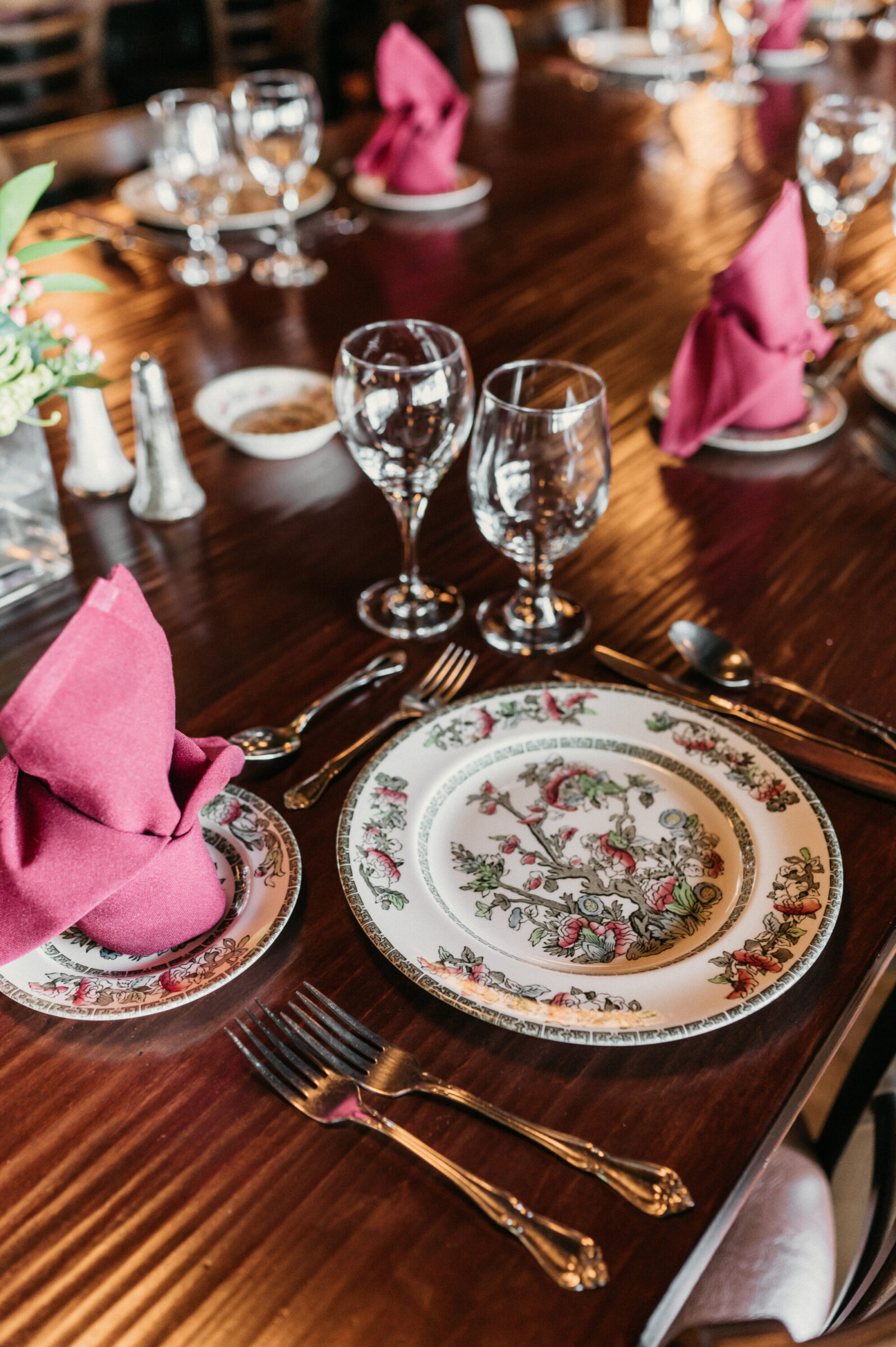 Table setting with floral plates, pink napkins, and wine glasses.