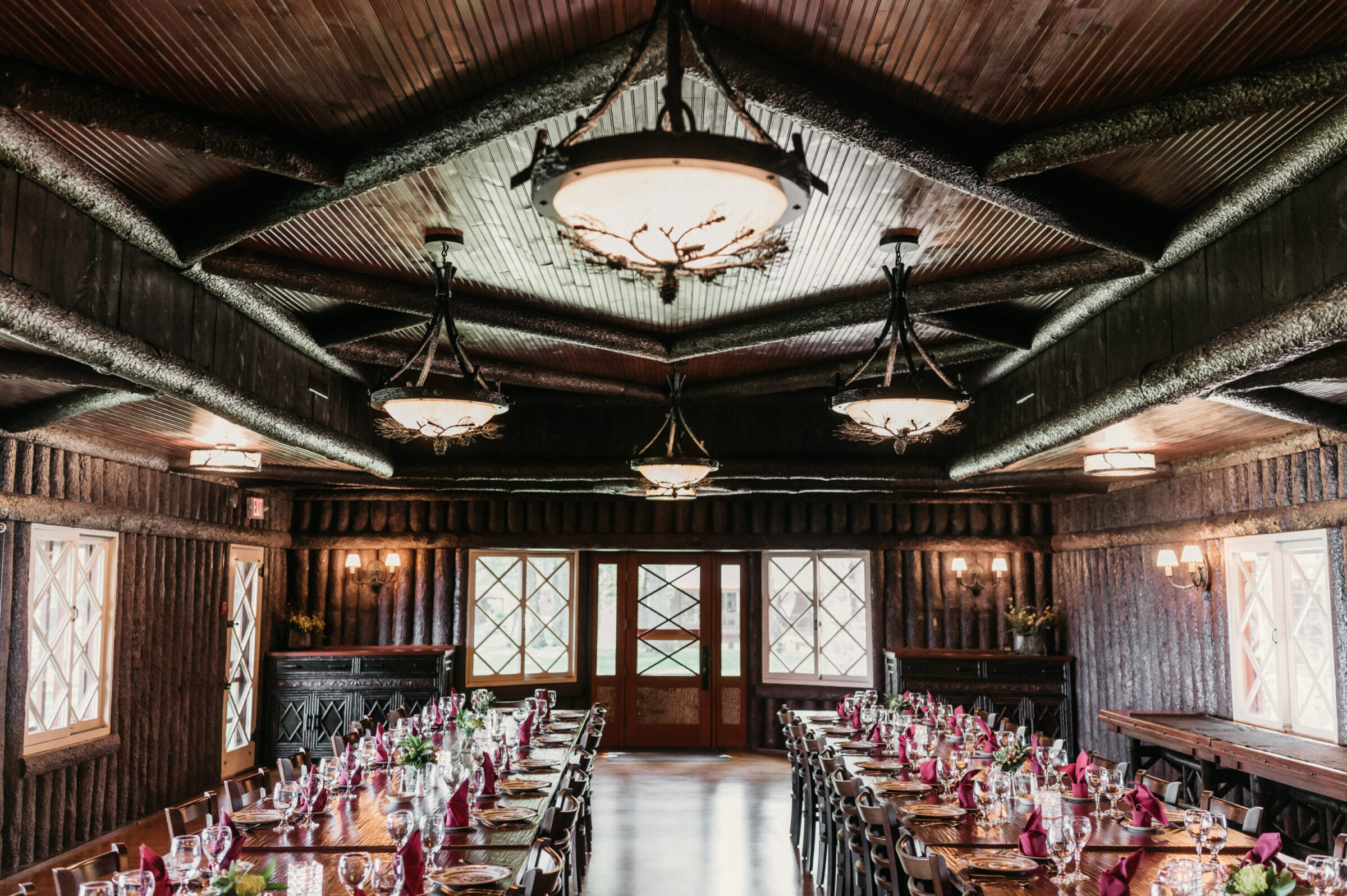 Rustic lodge dining room with long tables set for a meal, featuring log walls and ornate chandeliers.