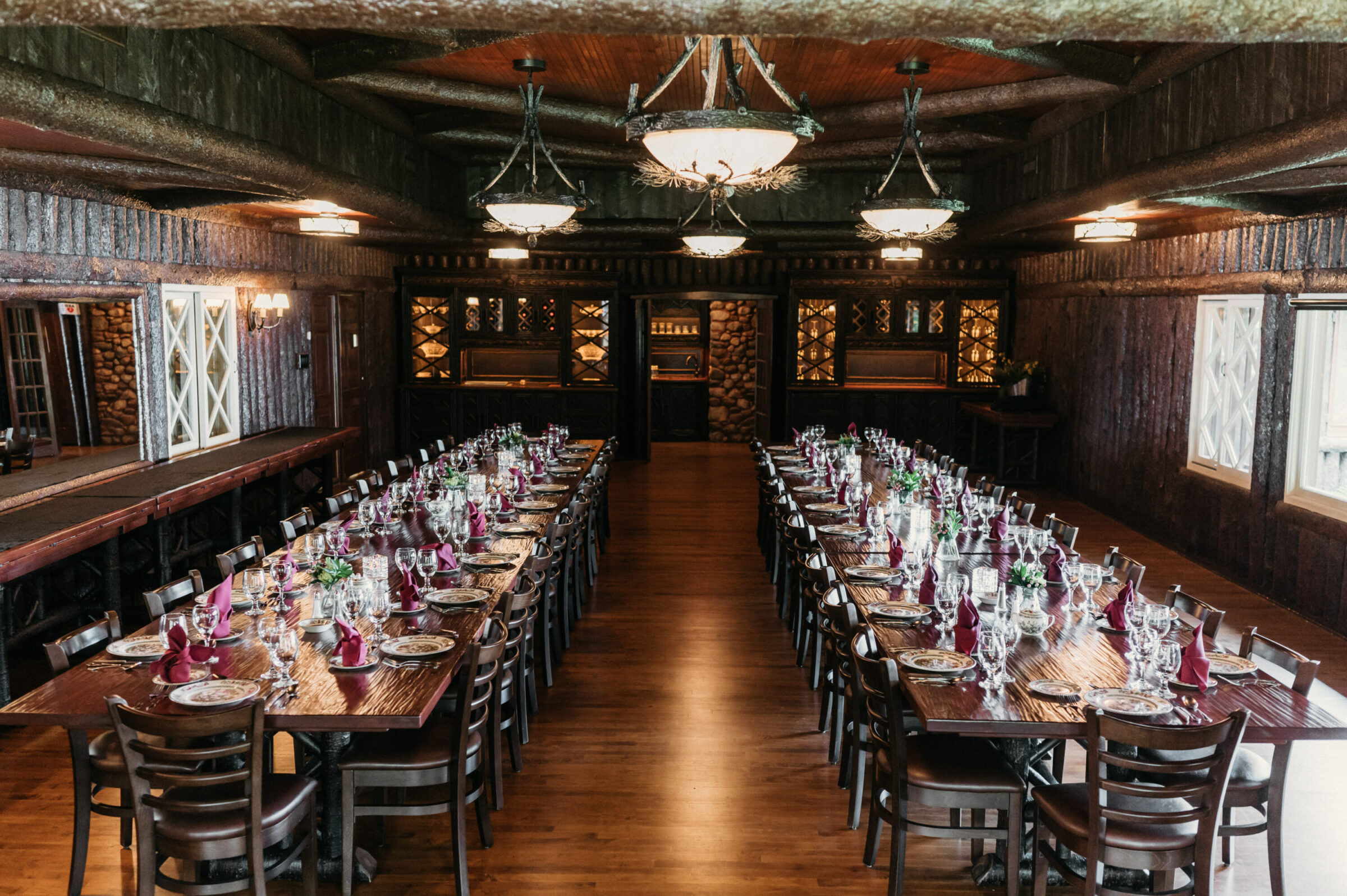 Two long dining tables set with plates, glasses, and burgundy napkins in a rustic room.