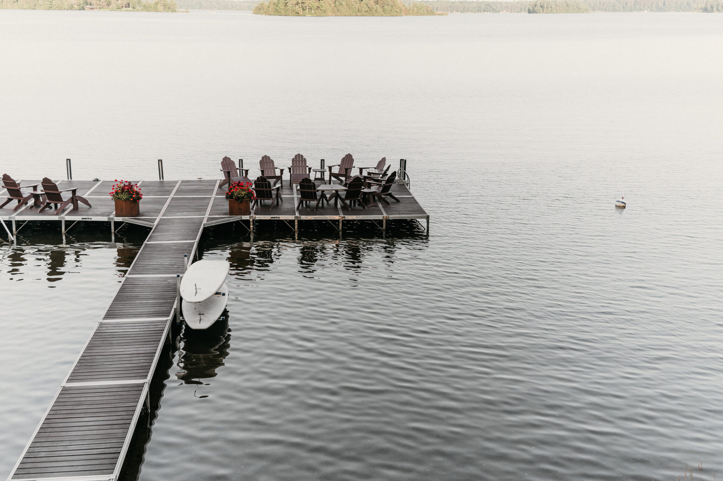 Dock with Adirondack chairs, tables, and a surfboard on a calm lake.