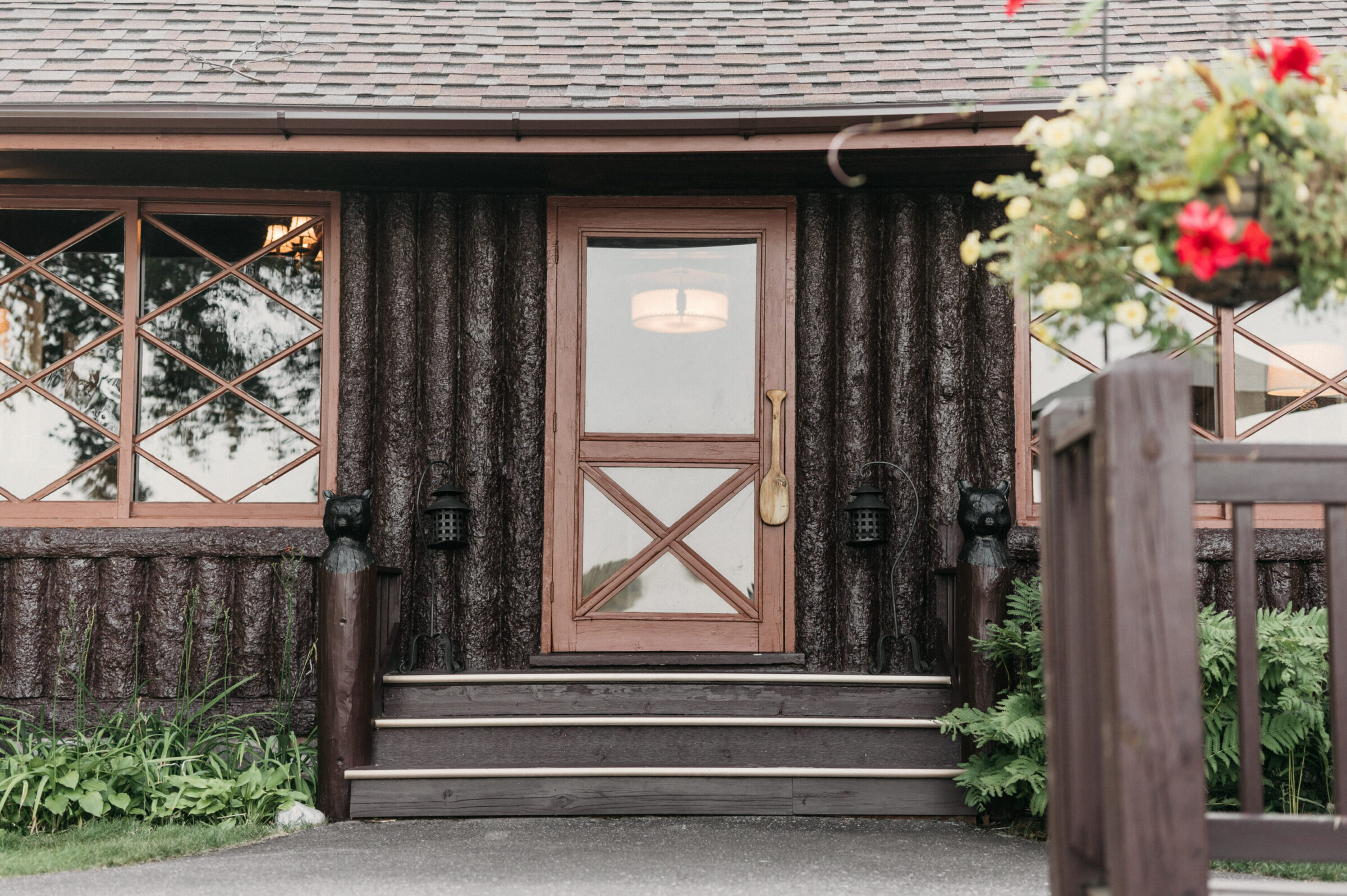 Log cabin entrance with wooden door, diamond-paned window, and hanging flowers.