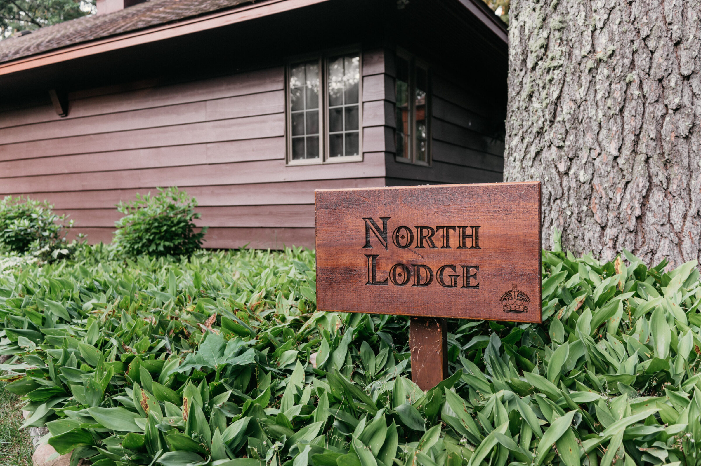 Wooden sign reading "North Lodge" in front of a brown building and green foliage.