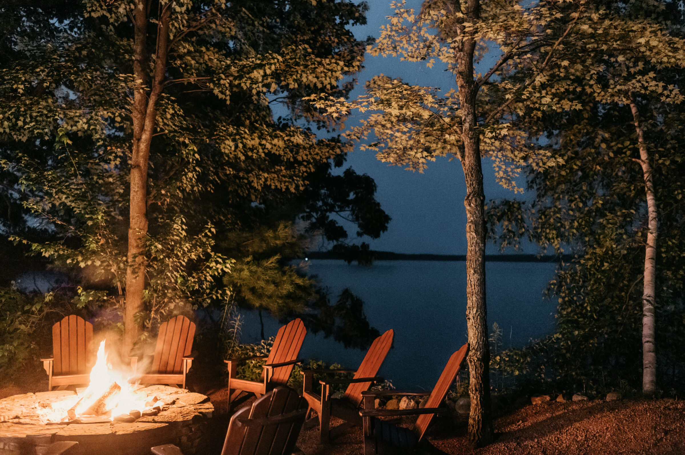 Campfire with Adirondack chairs by a lake at dusk.
