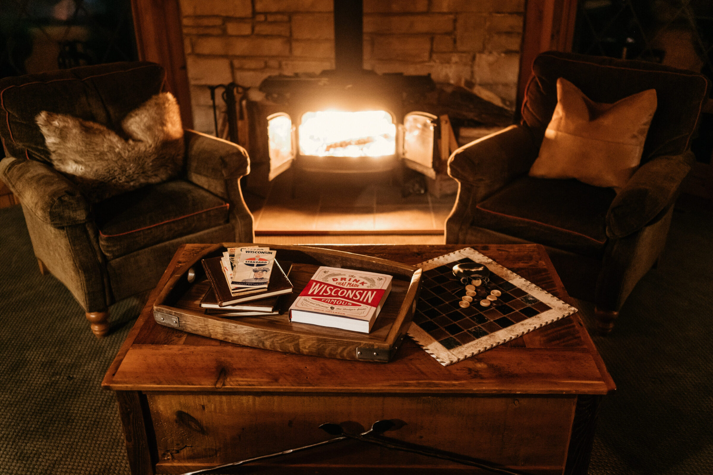 Cozy chairs by a fireplace with a tray of books and a checkerboard on a wooden table.