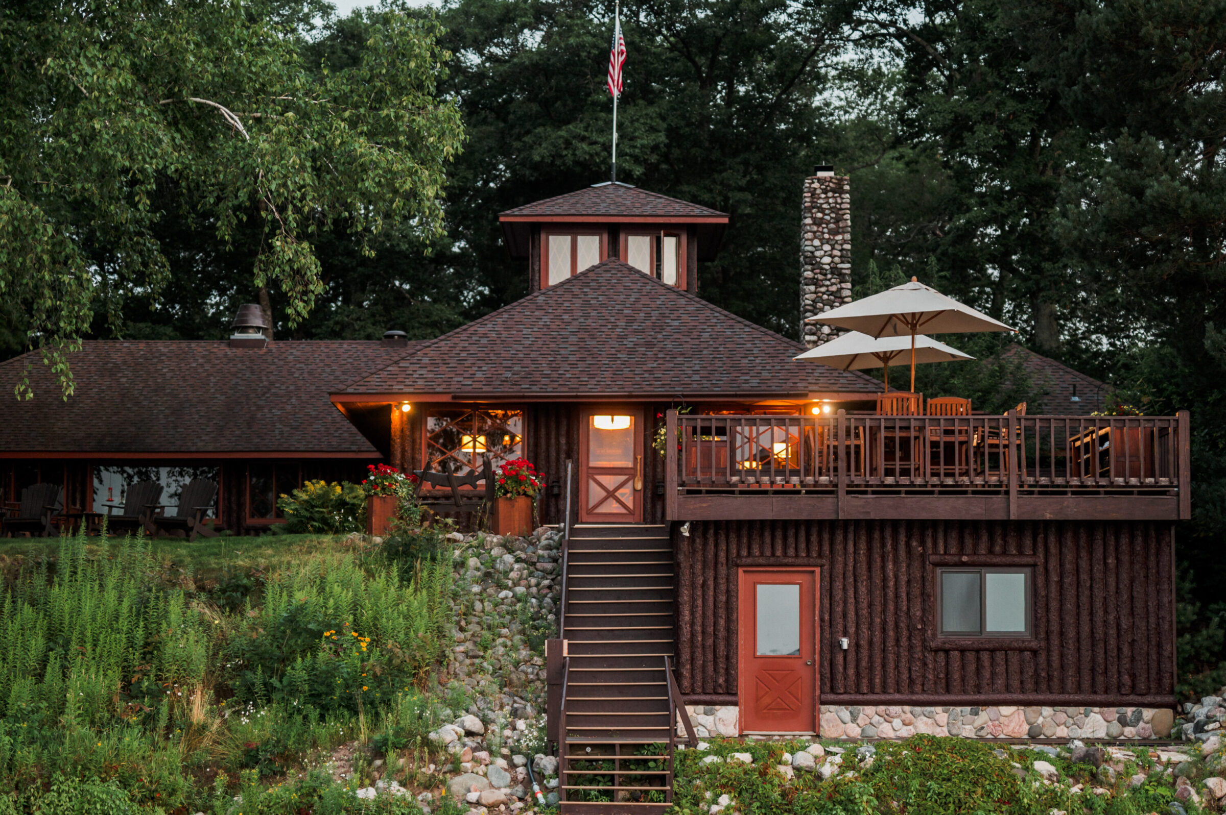 Log cabin with a deck, stone chimney, and American flag on a pole.