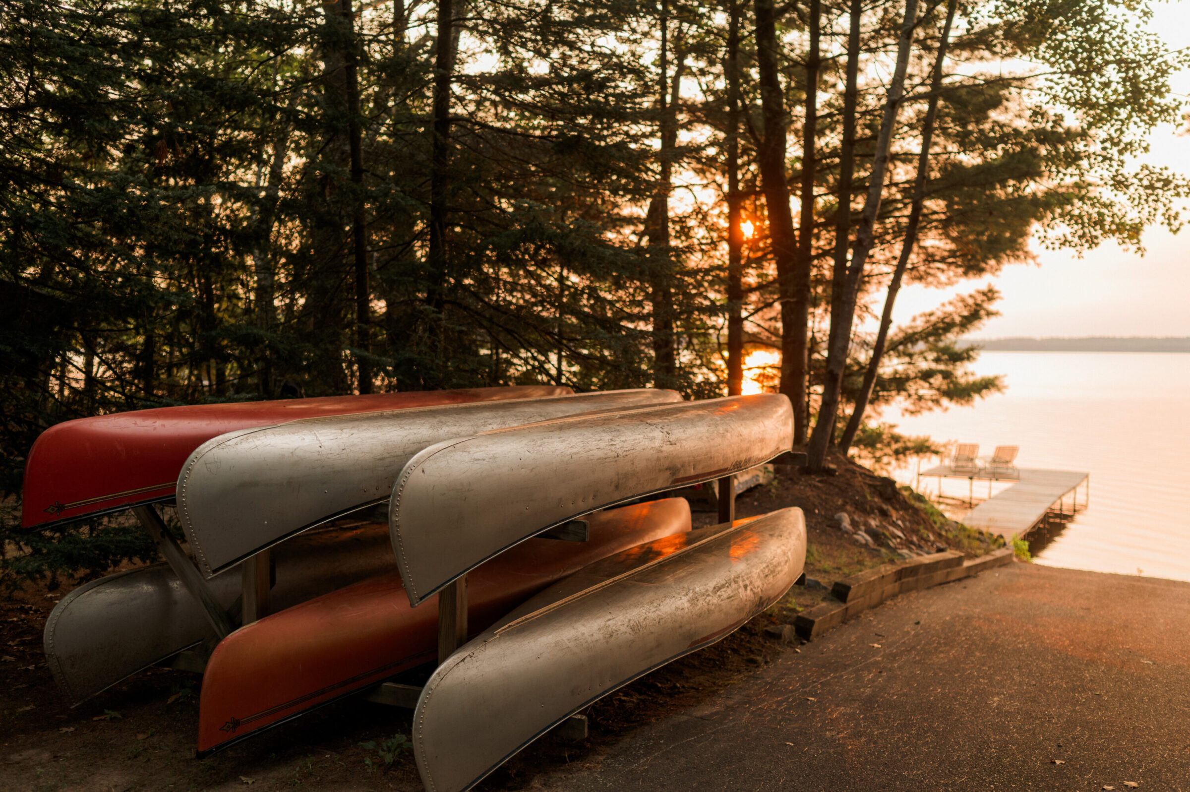 Canoes stacked on a rack by a lake at sunset, with a dock and chairs in the distance.