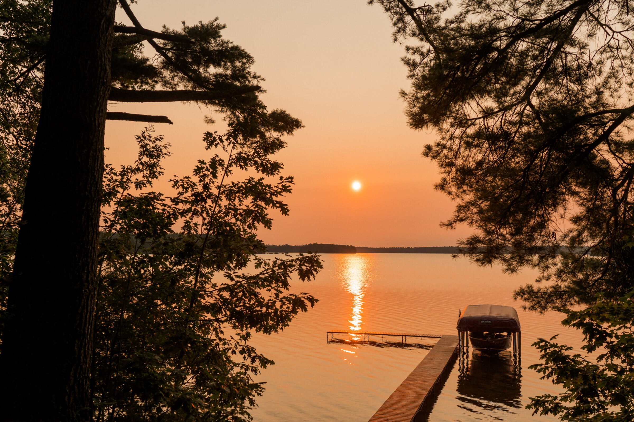 Sunset over a calm lake with a boat dock and trees framing the view.