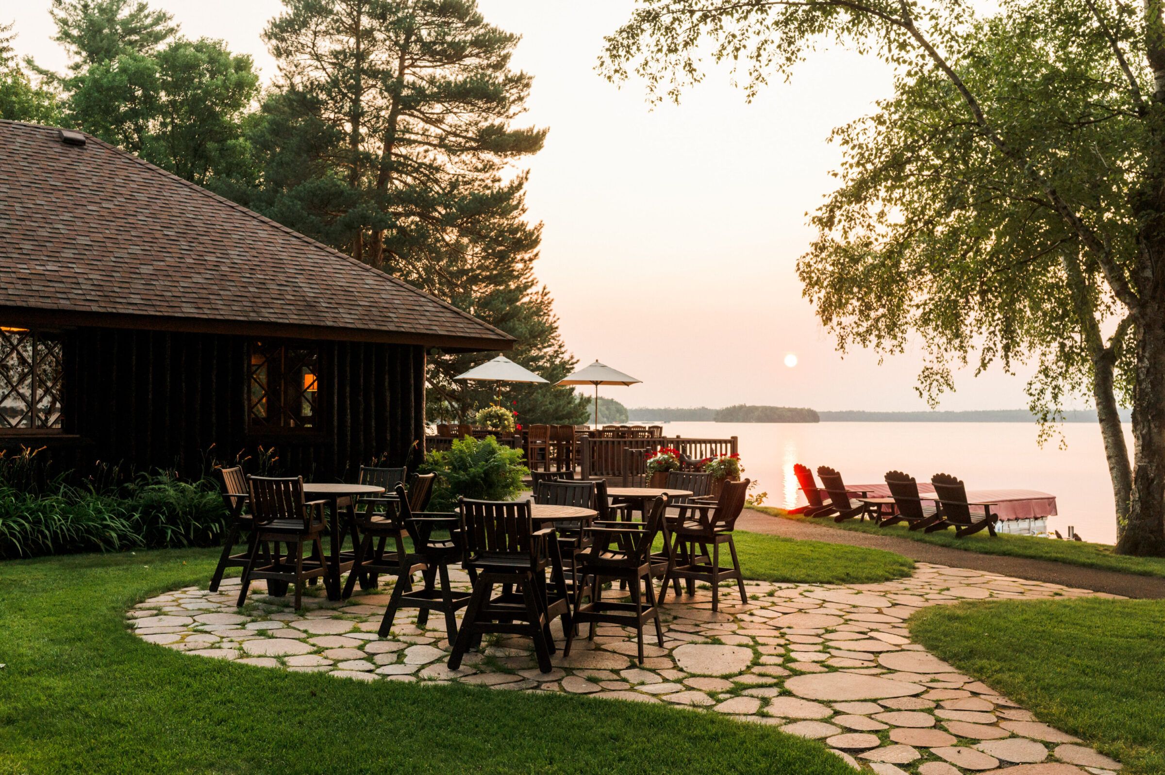 Outdoor seating area with tables and chairs on a stone patio overlooking a lake at sunset.
