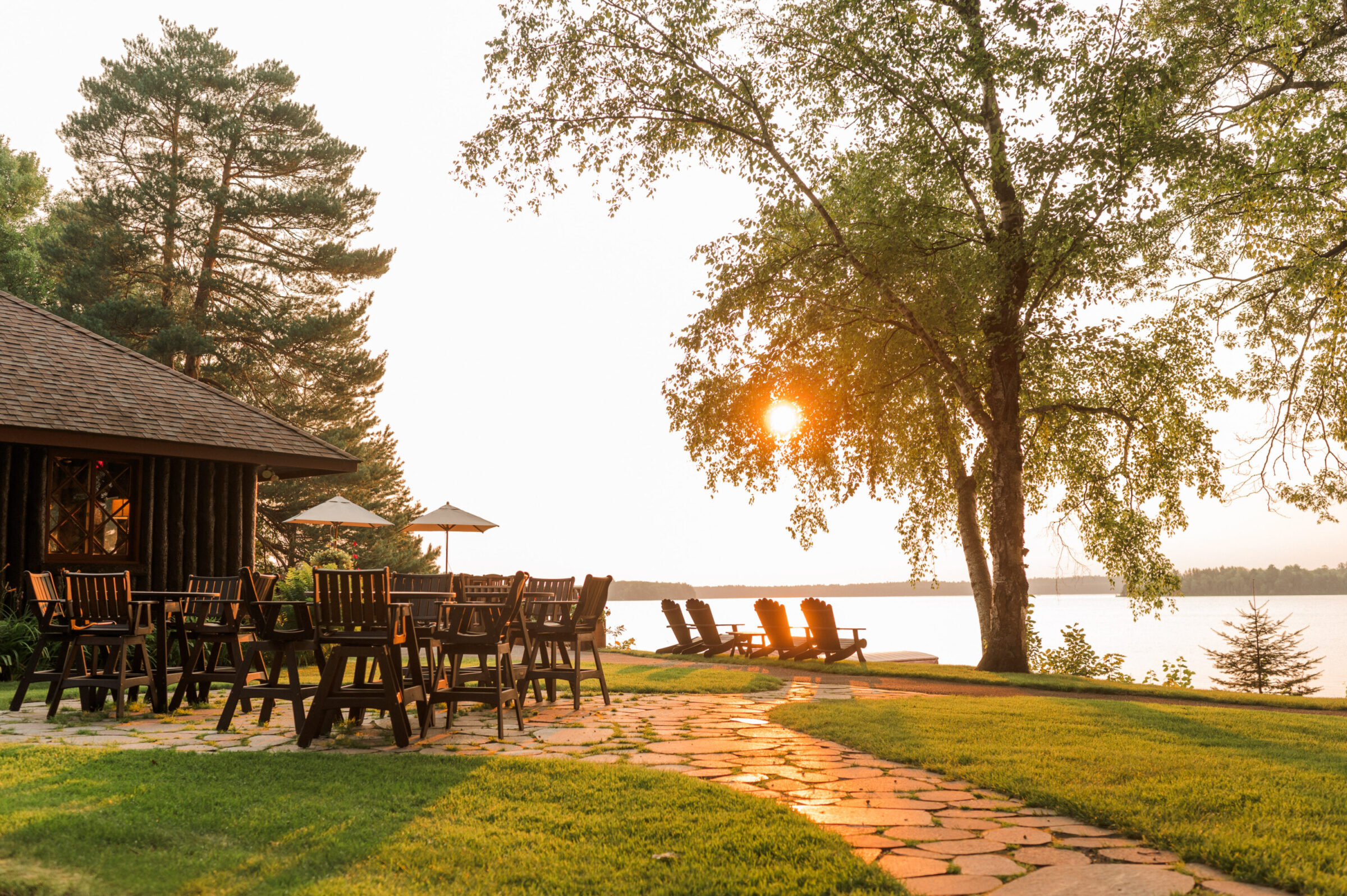 Lakeside patio with wooden tables, chairs, and Adirondack chairs at sunset.