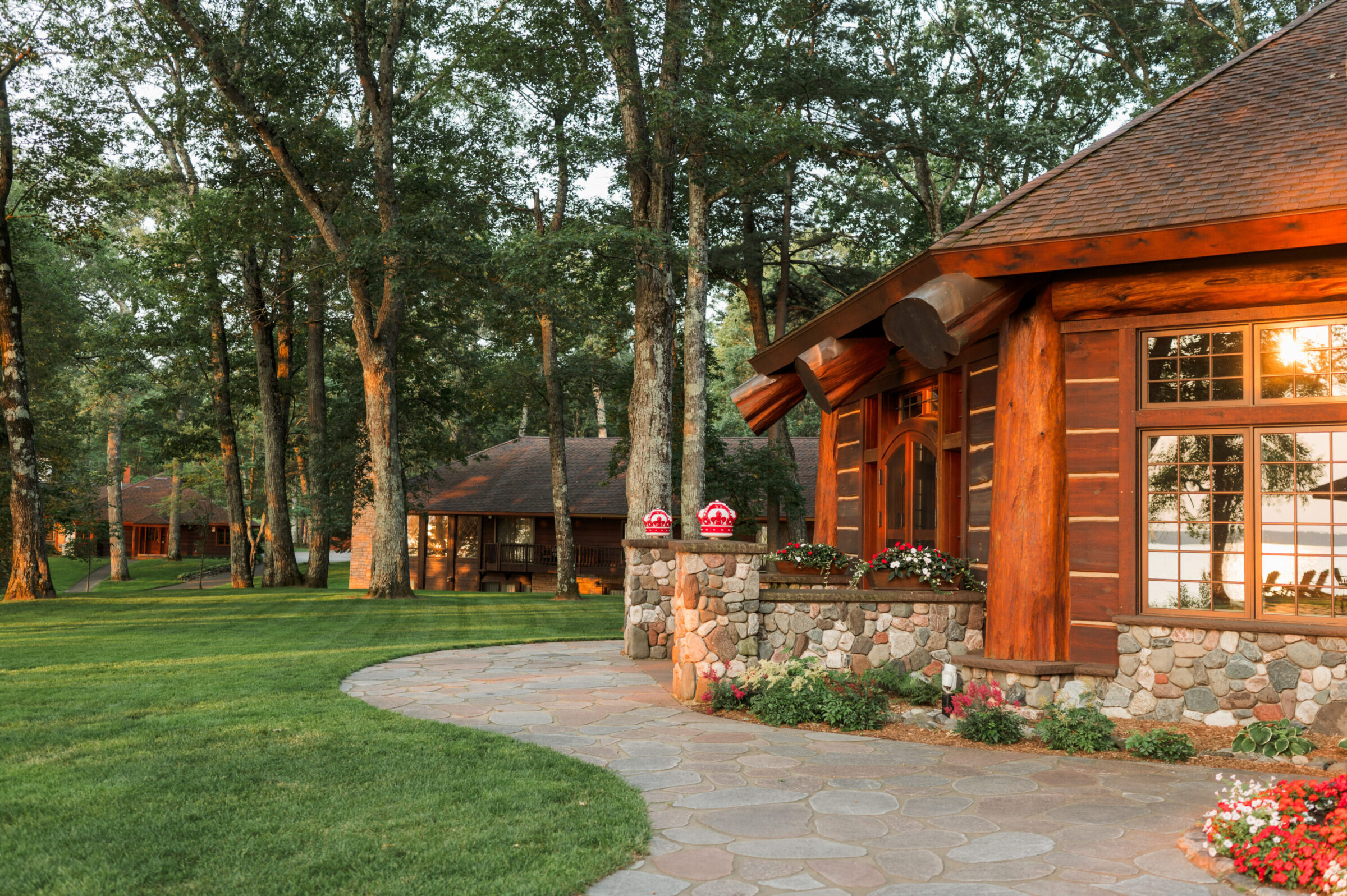 Log cabin with stone accents and flowers, surrounded by trees and a lake.