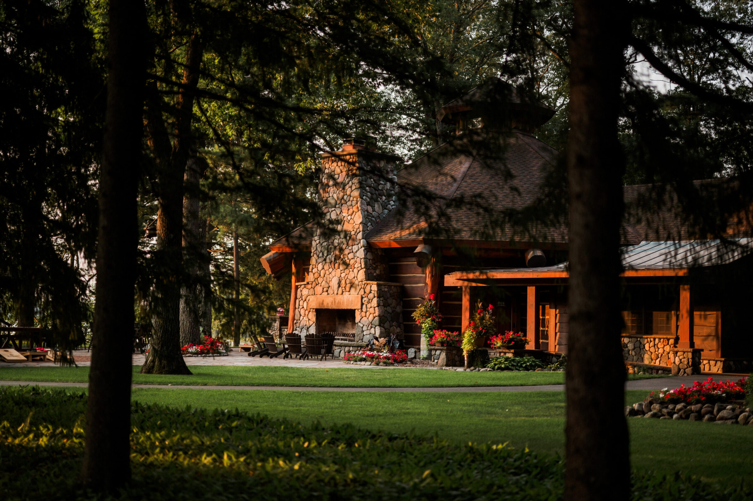 Log cabin with stone fireplace and porch, surrounded by trees and flowers.