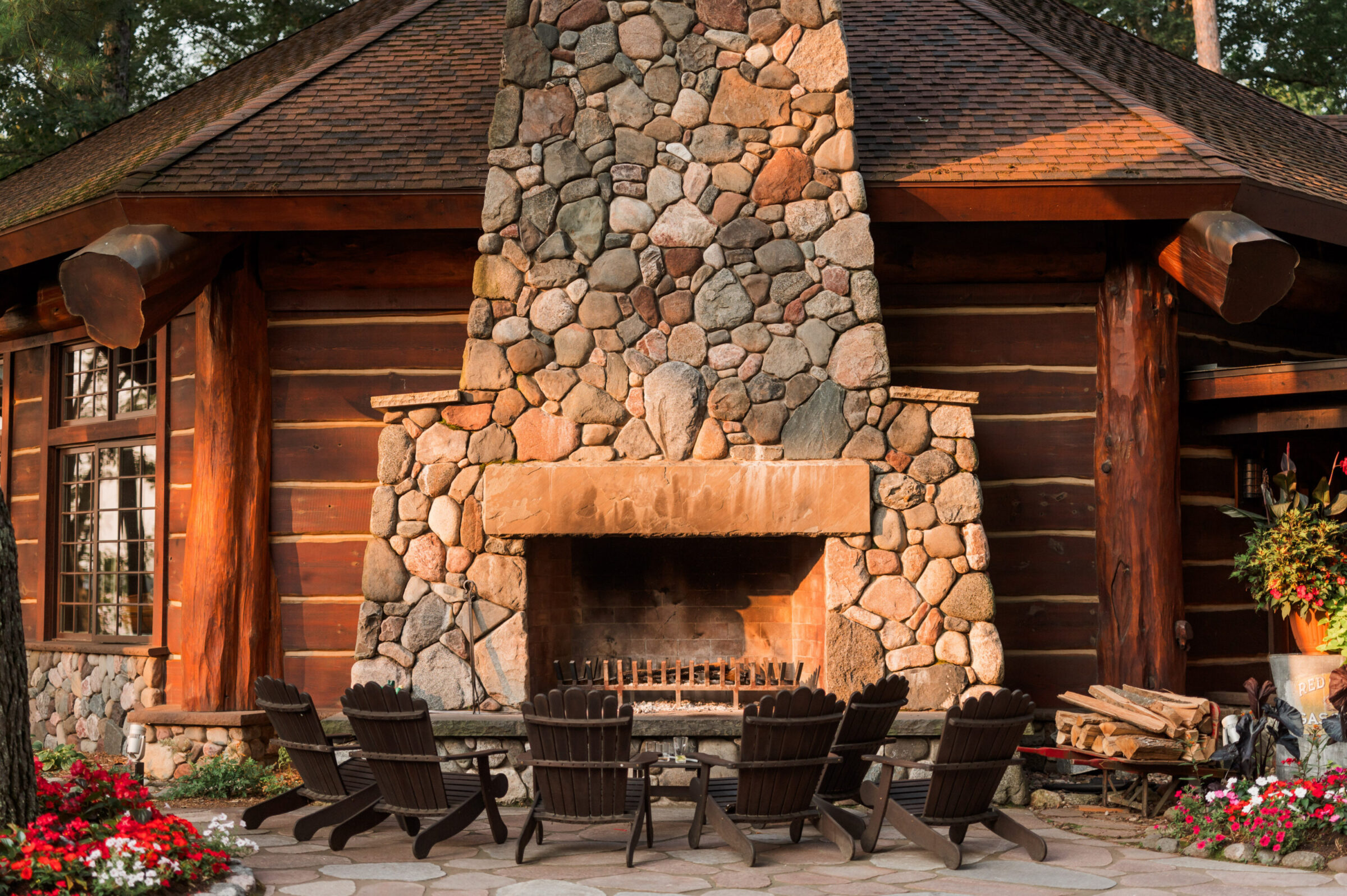 Log cabin with large stone fireplace and Adirondack chairs on patio.